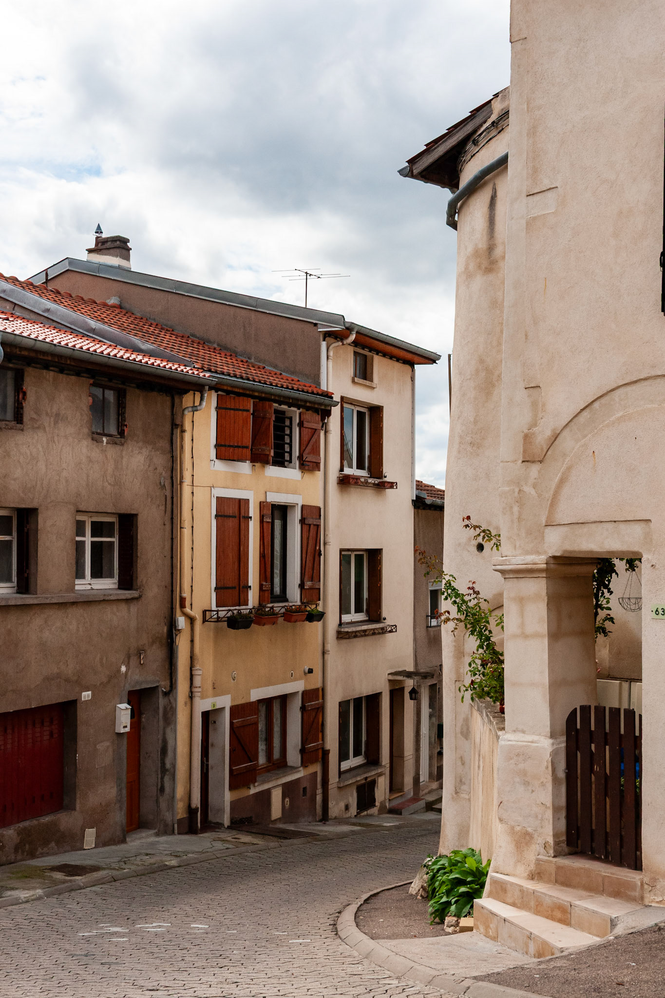 A quiet, winding cobblestone street in a historic French town, featuring multi-story residential buildings with traditional wooden shutters and terracotta tile roofs under a soft, cloudy sky.