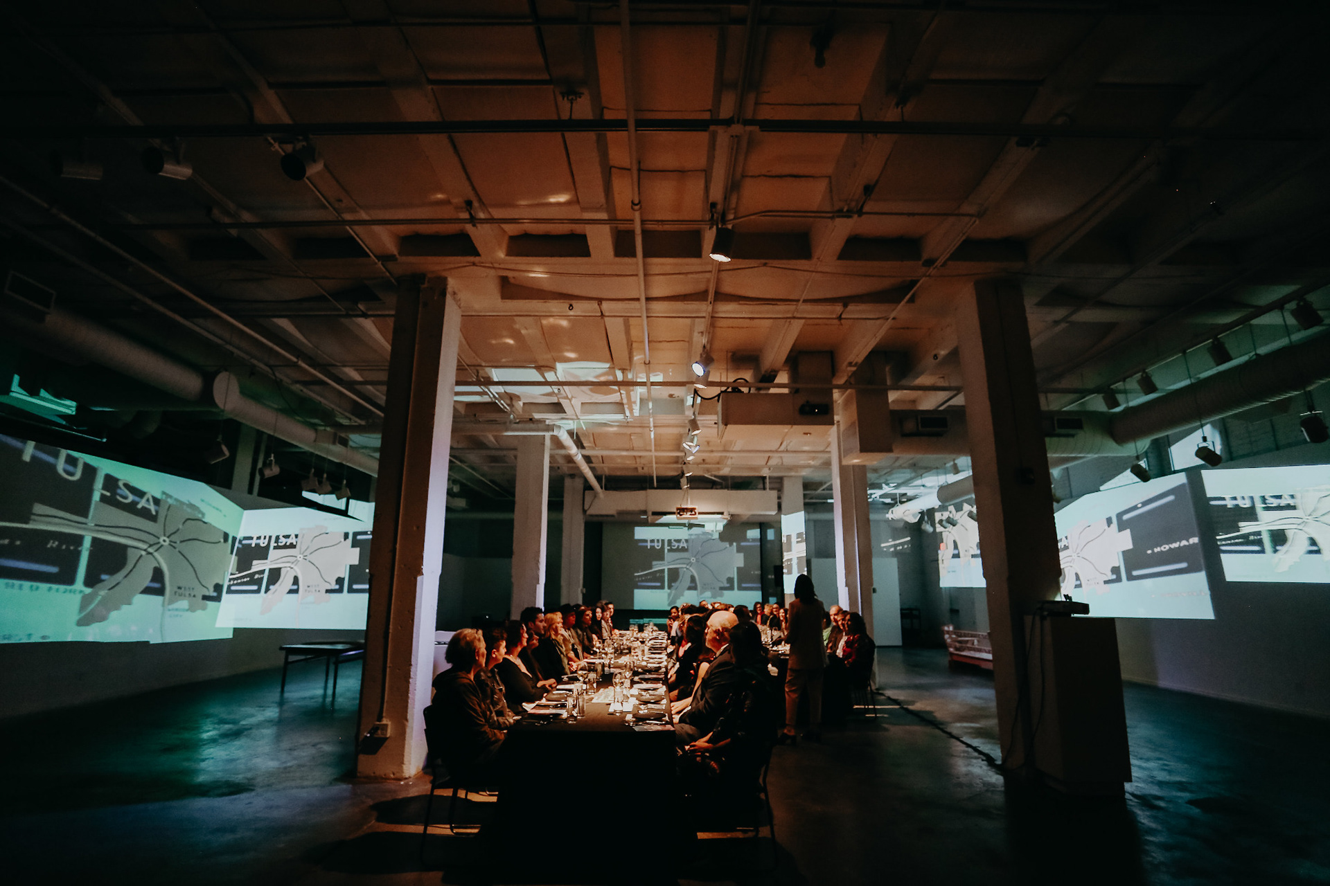 Guests seated at a long dinner table surrounded by large-scale video projections in an industrial gallery.