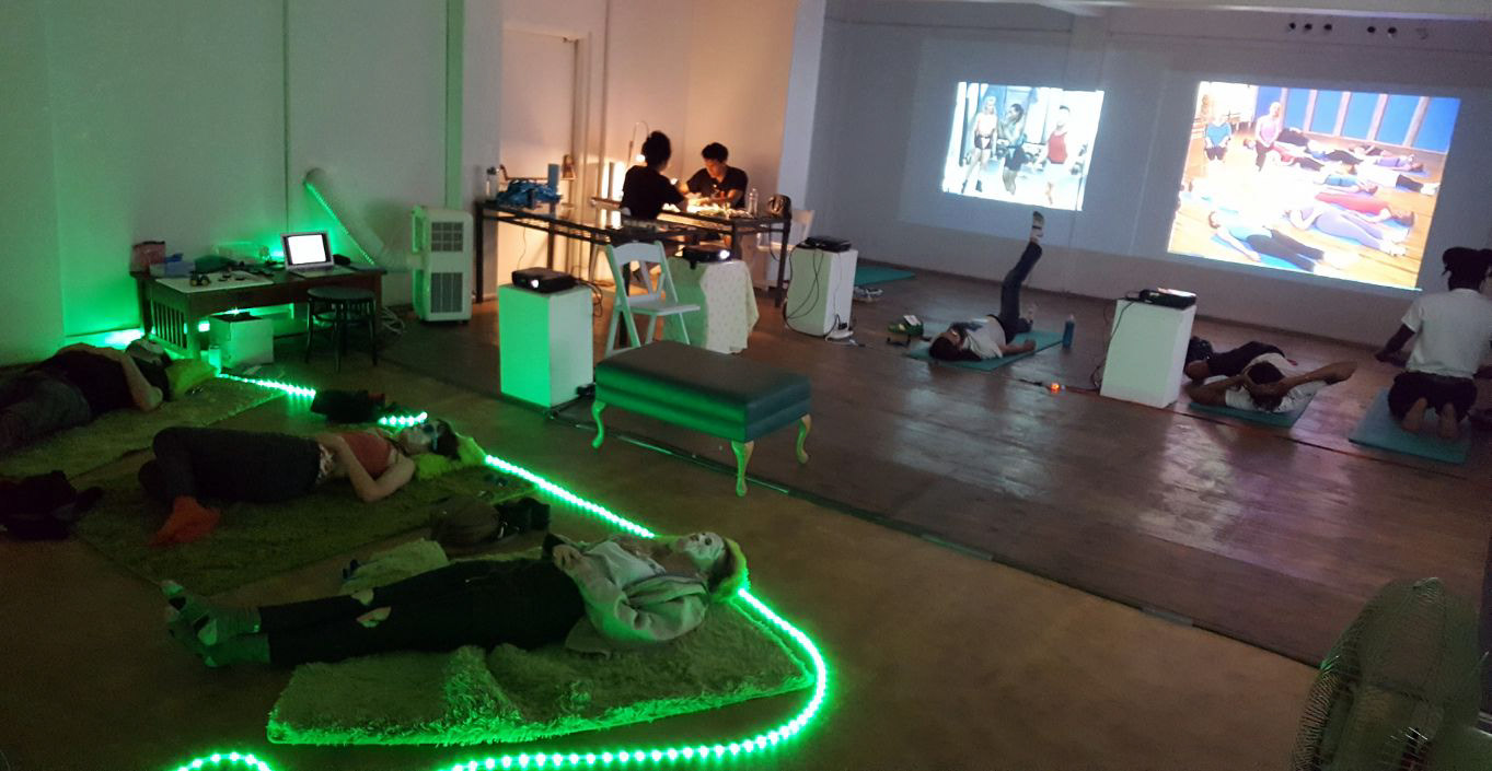 Visitors reclining on rugs and yoga mats inside Video Lounge and Spa, with green floor lighting and projected videos, San Diego, 2019.