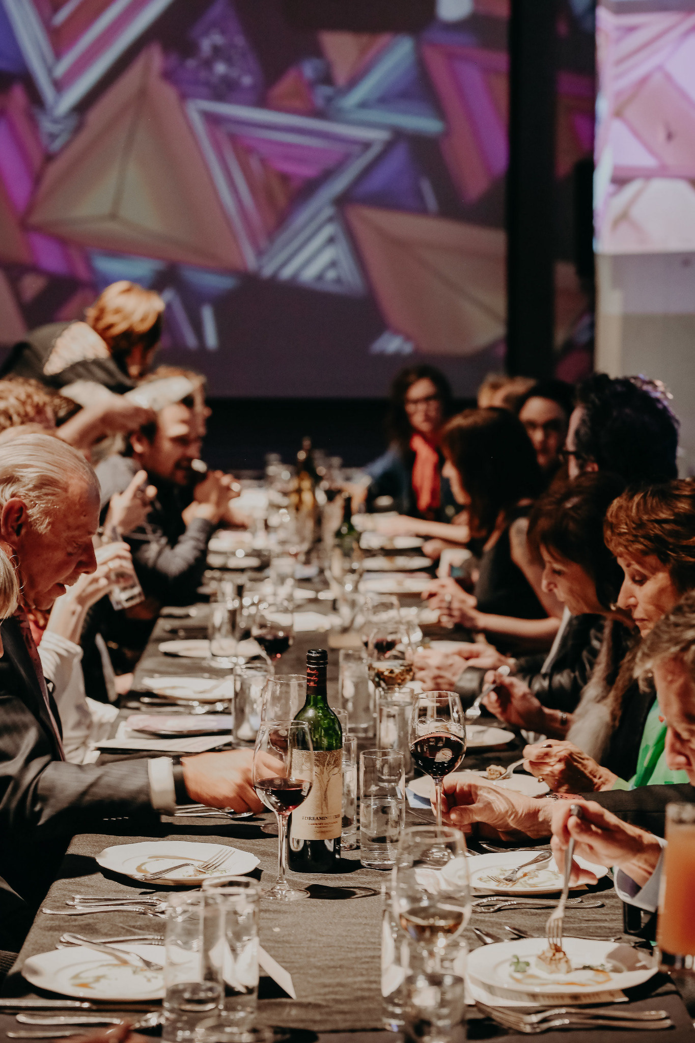 A close view down a communal dinner table, with guests eating beneath a colorful projected image.