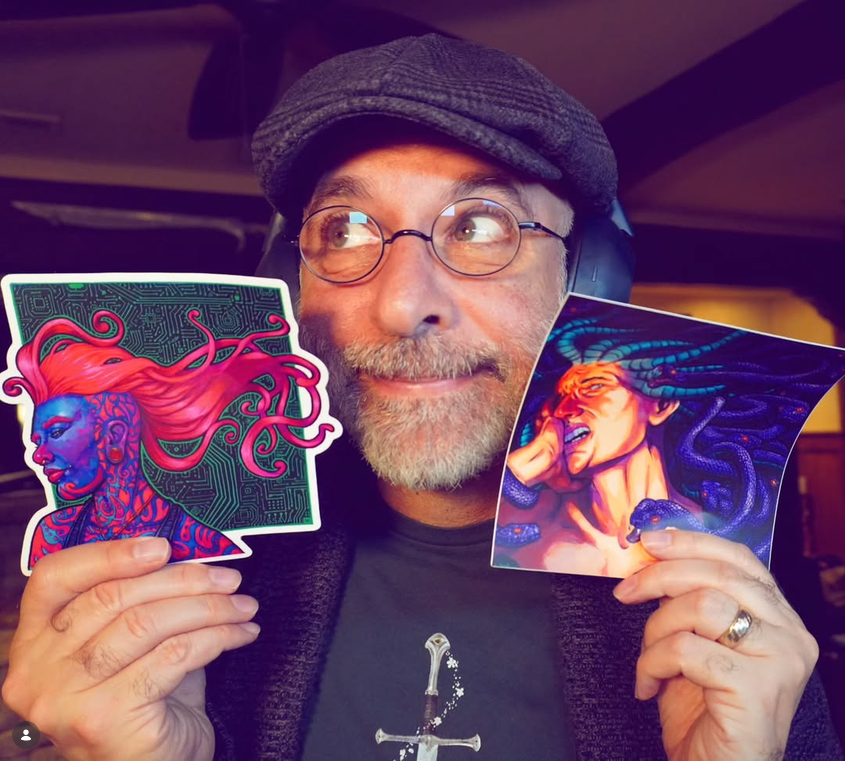Artist Scott Christian Sava, wearing round glasses and a gray flat cap, holds two colorful fantasy portraits beside his face: a blue-skinned figure with flowing red hair and a snake-haired Medusa.
