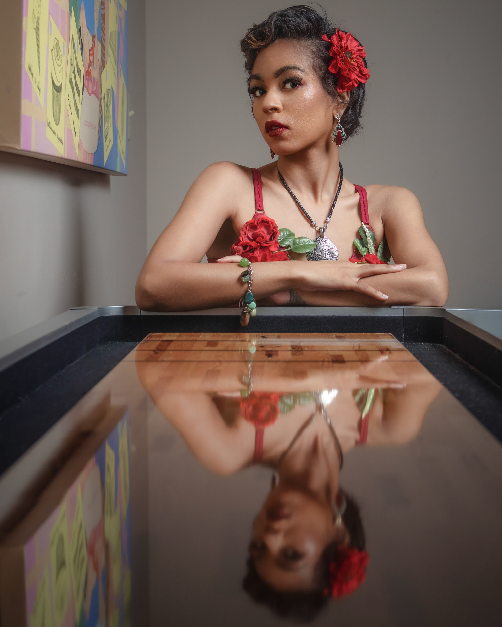 A striking portrait of a woman adorned with vibrant floral accessories, leaning on a reflective surface that creates a mirrored effect. The composition is enhanced by an abstract painting in the background, contributing to a mood of artistic elegance and confident beauty.