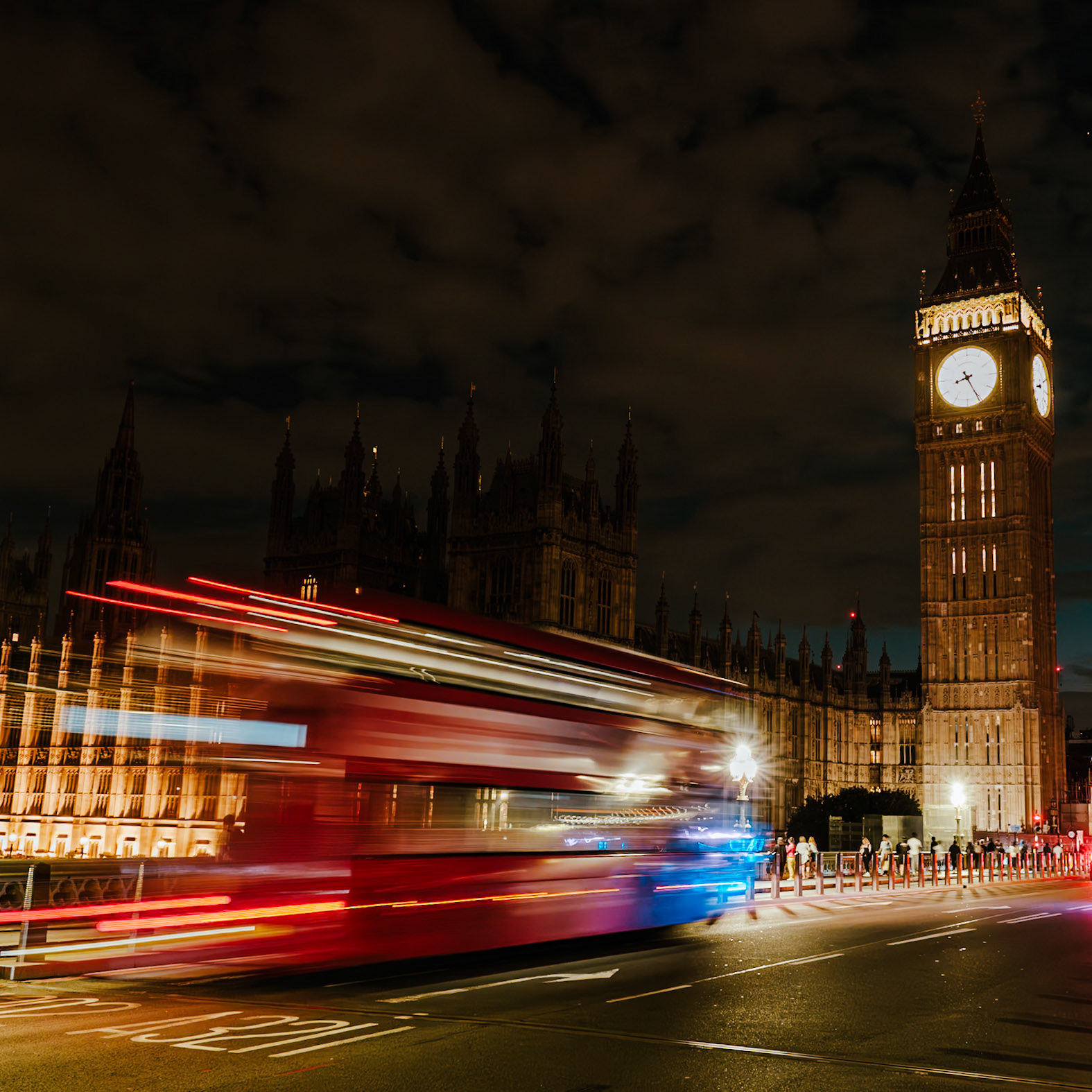 A long-exposure view of London’s Elizabeth Tower and the Houses of Parliament catches red double‑decker buses streaking past at night, contrasting historic stonework with modern motion.