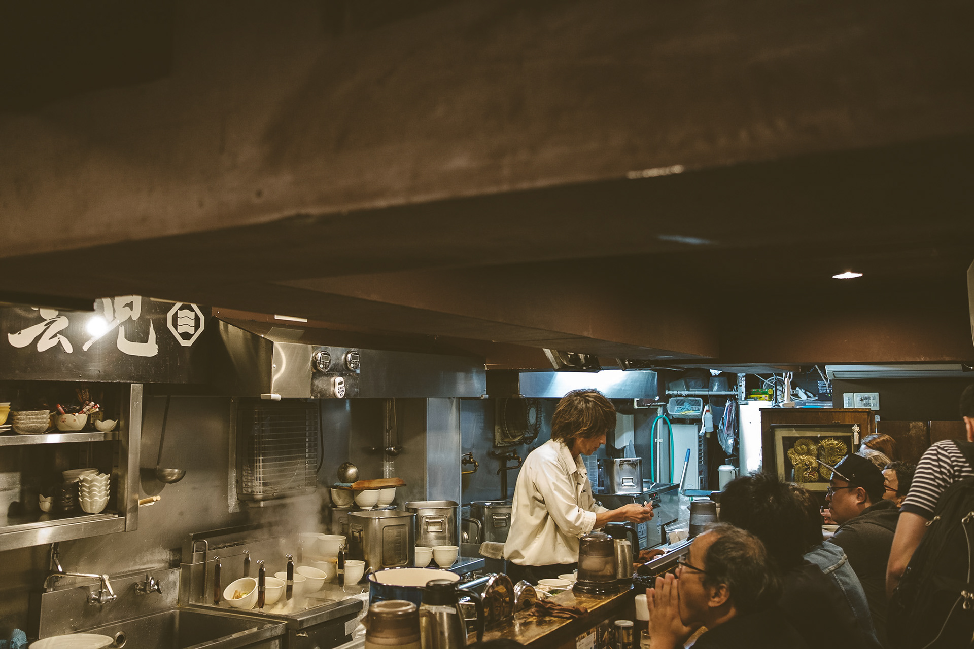 Chef preparing ramen bowls in a small Tokyo shop interior, with steam rising and customers waiting at the counter.