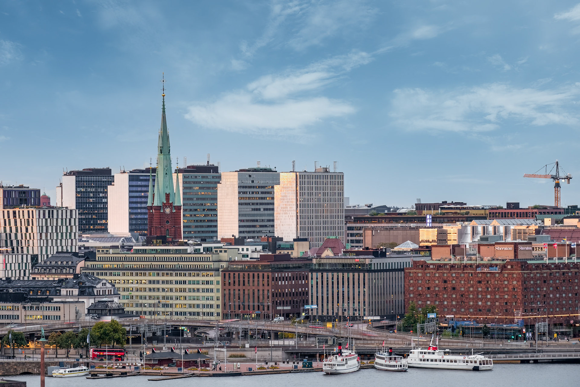 The slender copper spire of Stockholm’s Klara Kyrka pierces the skyline amid modern offices along Riddarfjärden, captured on a clear late‑afternoon day.