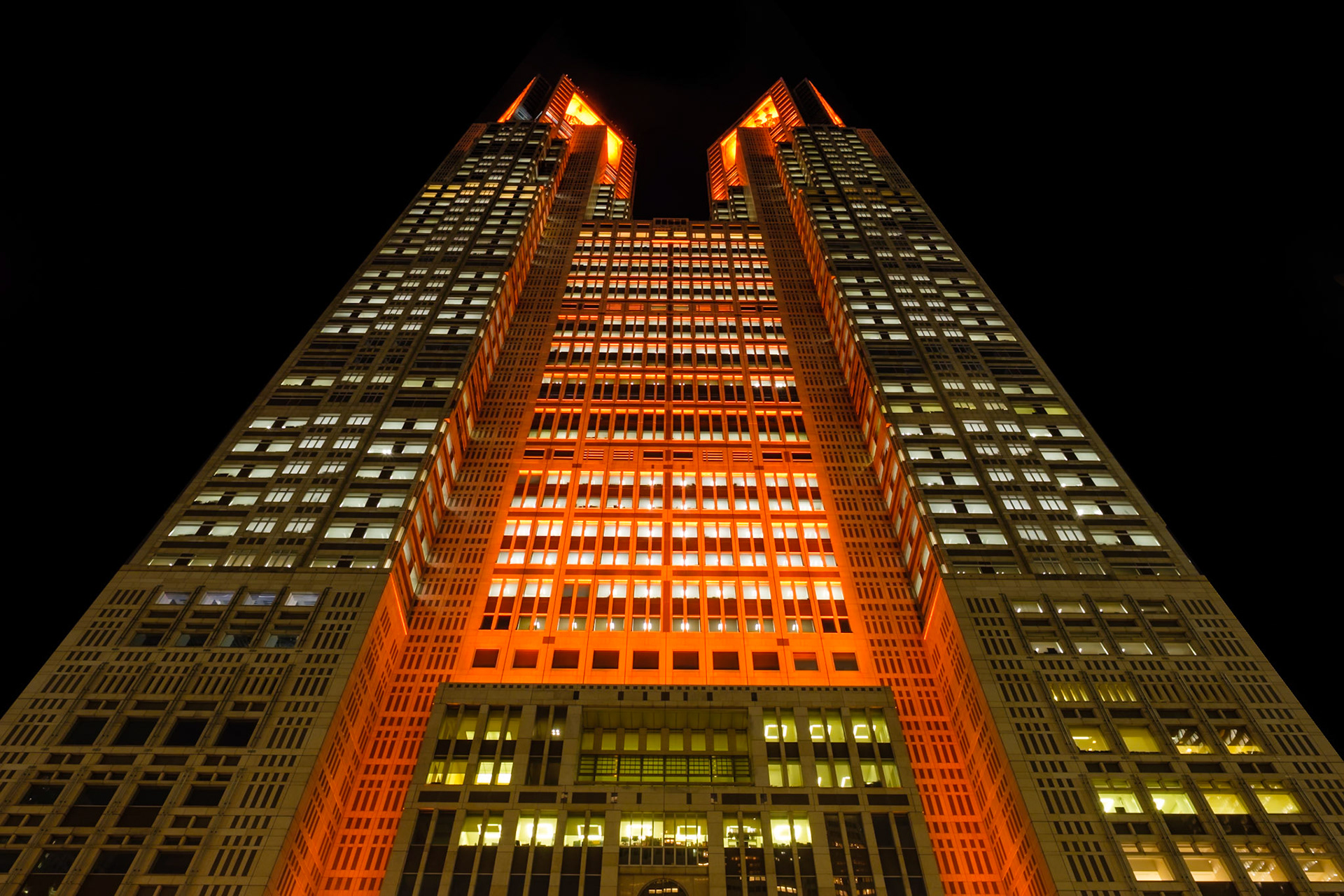 Bathed in vivid orange lights, the twin towers of the Tokyo Metropolitan Government Building soar into the night sky, epitomizing Shinjuku’s modern skyline.
