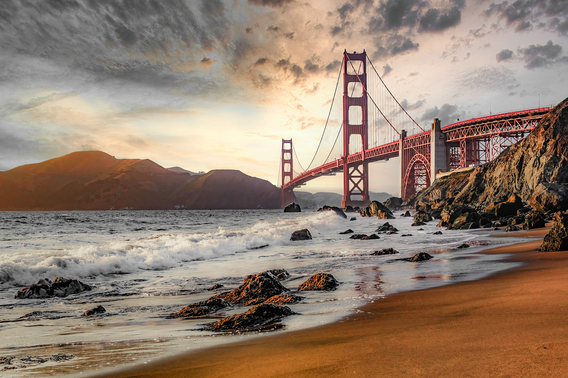 Waves lap the sands of Marshall’s Beach as the Golden Gate Bridge blazes red against a pastel sunset over San Francisco Bay and the Marin Headlands.