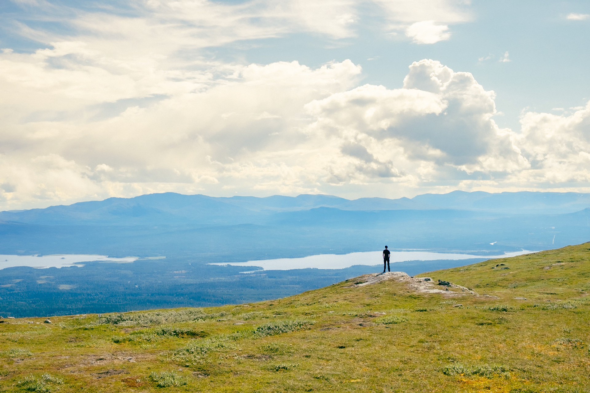 A lone hiker surveys the sweeping vistas of Åre’s mountain lakes and rolling Jämtland highlands from the windswept summit of Välliste under billowing summer clouds.