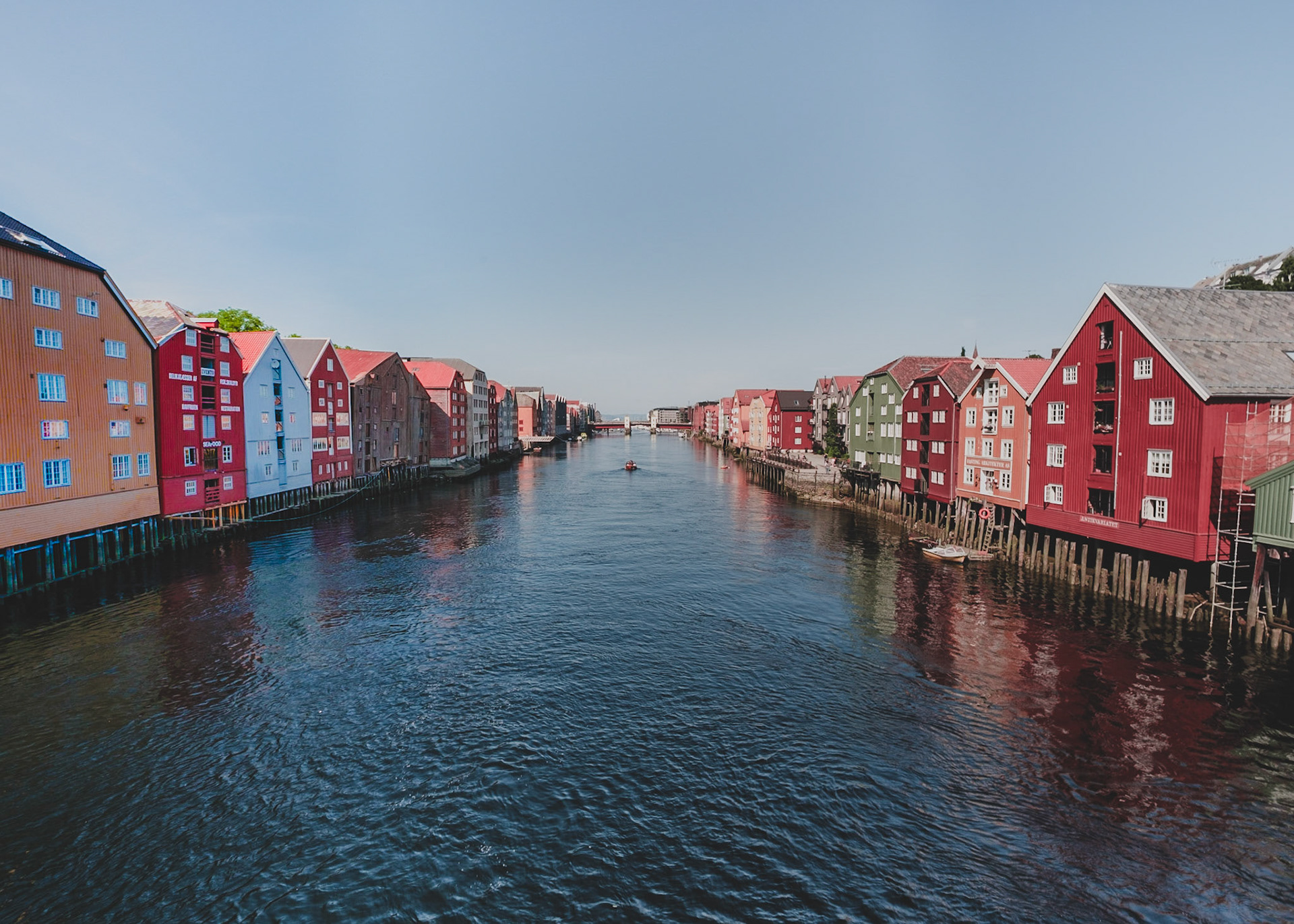 The historic wharf houses of Bakklandet in Trondheim line the Nidelva River, their red and pastel facades mirrored in the calm water beneath a cloudless Nordic sky.