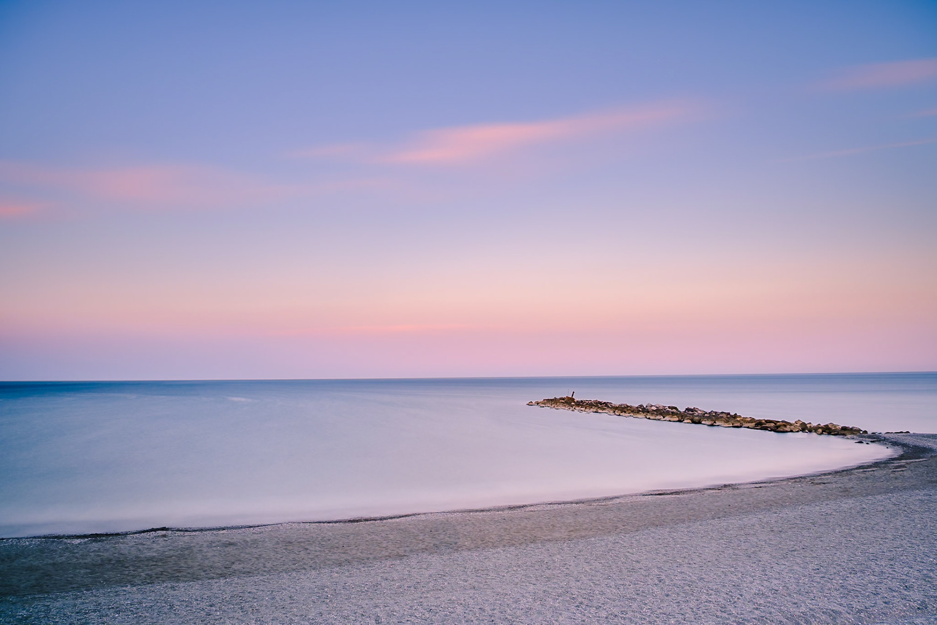 A smooth Mediterranian Sea meets the pastel evening sky off Spain Goald coast, where a stone breakwater extends into the tranquil horizon.