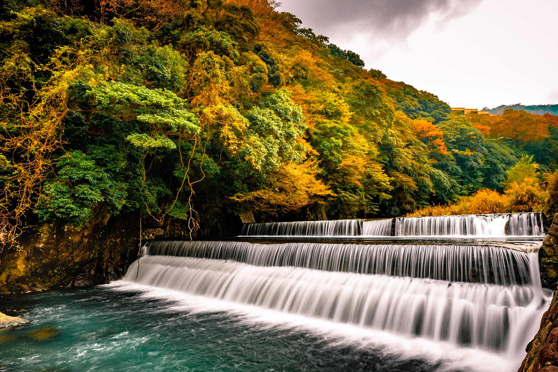 Layered waterfall amid autumn foliage, where turquoise waters flow through emerald and golden leaves in rural Japan.