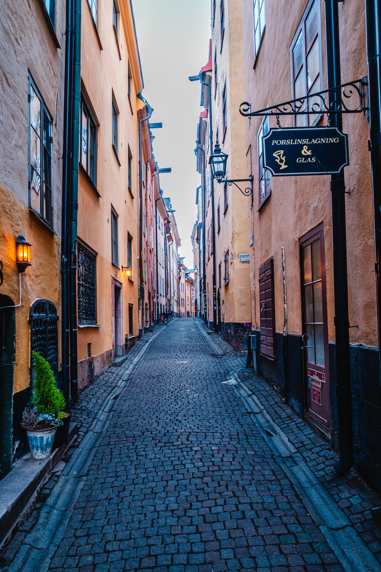 Cobblestone alley in Stockholm’s Old Town (Gamla Stan), lined with yellow historic buildings, lanterns glowing, and a ‘Porslinslagning & Glas’ shop sign on Prästgatan