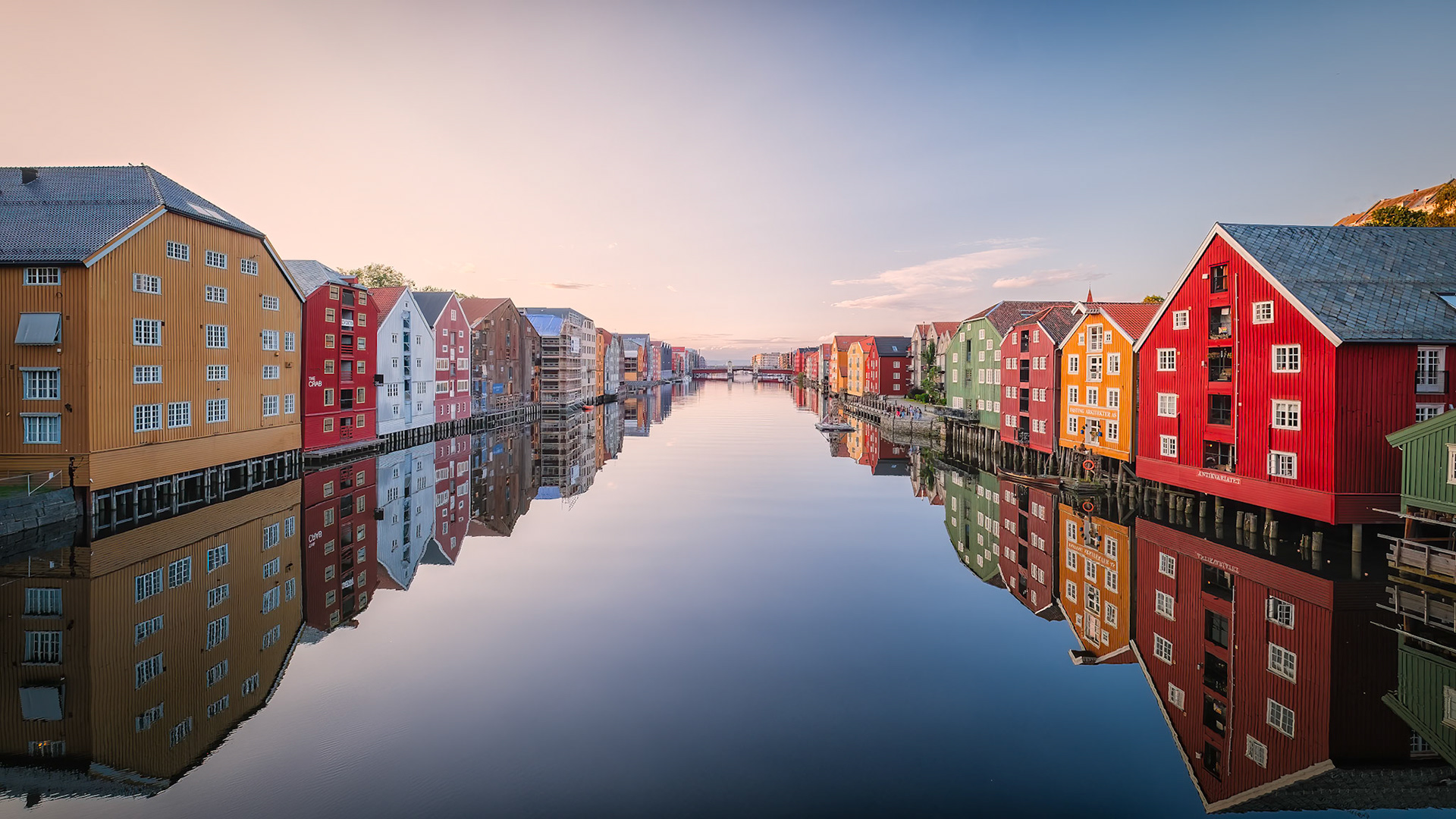 Colorful wooden warehouses of Trondheim’s Bakklandet district line the banks of the Nidelva River, their shuttered facades mirrored perfectly in the calm morning water.