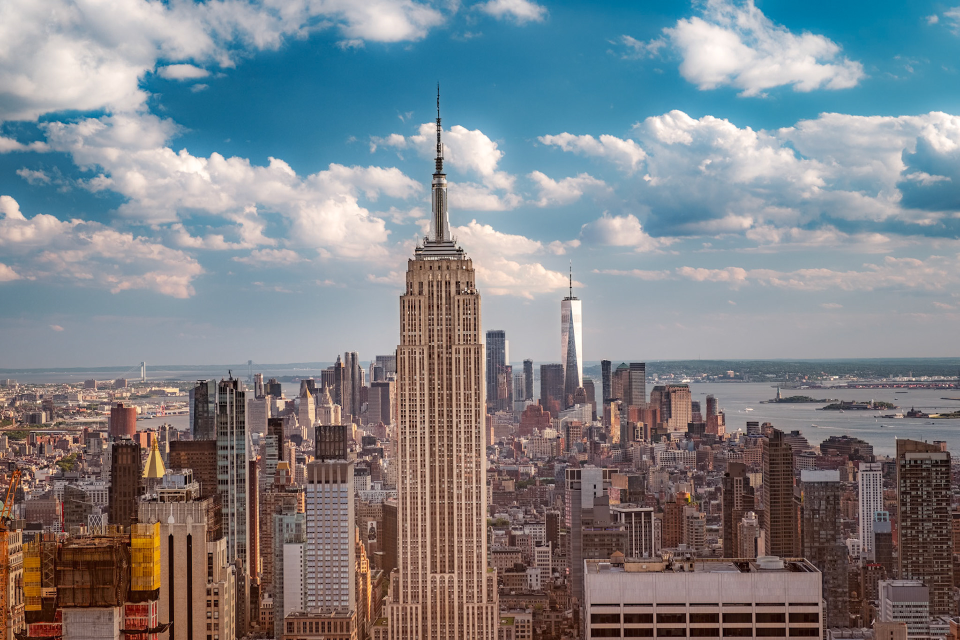 The Empire State Building towers over Midtown Manhattan while One World Trade Center and the rest of New York City stretch toward the harbor under a brilliant summer sky.