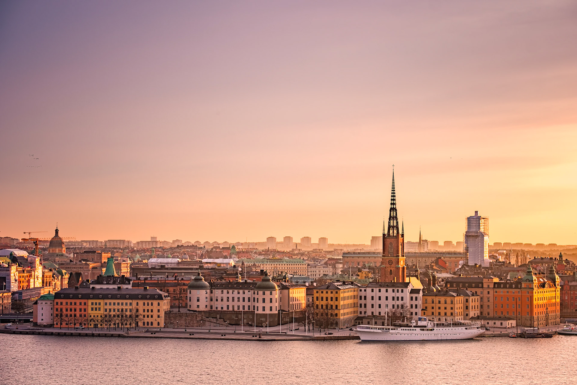 Stockholm’s Gamla Stan and Riddarholmen glow under a golden sunrise as the slender spire of Riddarholmskyrkan pierces the pastel sky above Riddarfjärden.