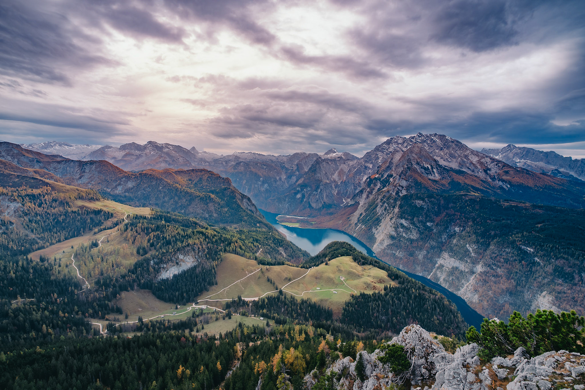 A sweeping panorama of Berchtesgaden National Park reveals the emerald waters of Lake Königssee cradled by the jagged Watzmann massif and rolling alpine meadows under gathering storm clouds.