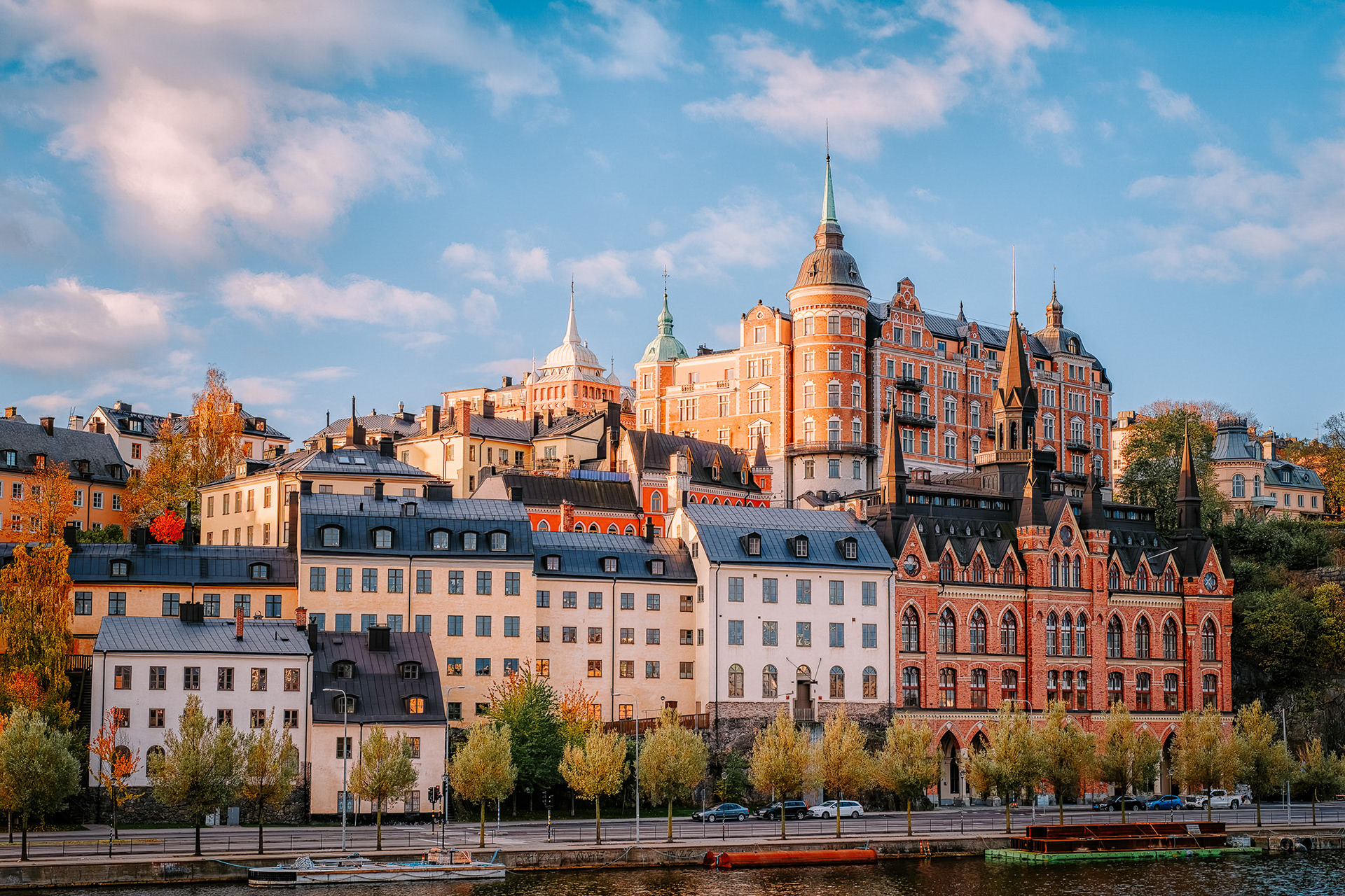 View from Munkbrohamnen in Stockholm showing historic buildings with colorful facades and distinctive turrets rising above Söder Mälarstrand, bathed in golden sunset light and reflected in the waters of Riddarfjärden
