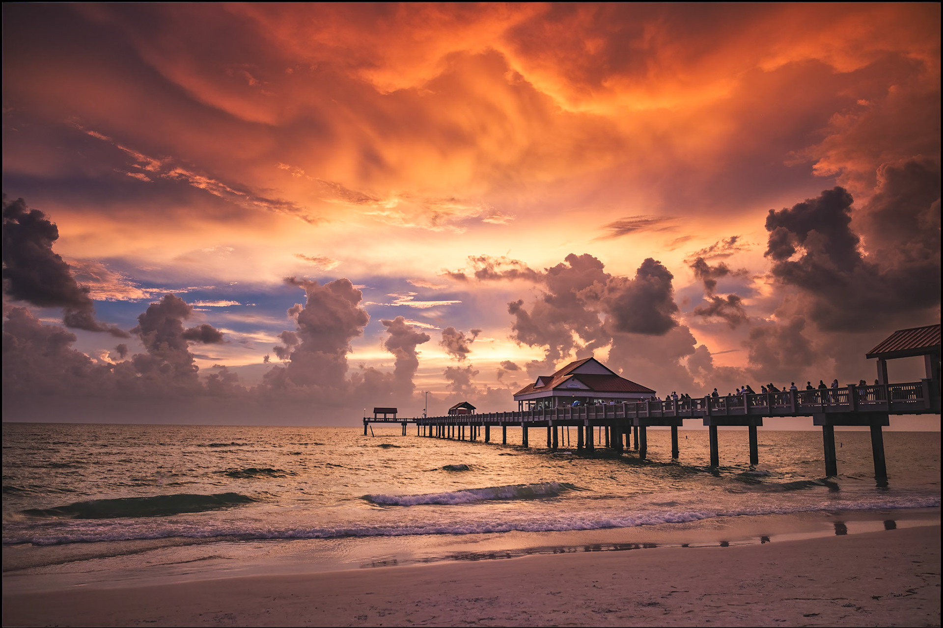 Fiery clouds ignite the sky above a tranquil Gulf beach as a wooden pier and pavilion reach into the sea, reflecting the glowing sunset.