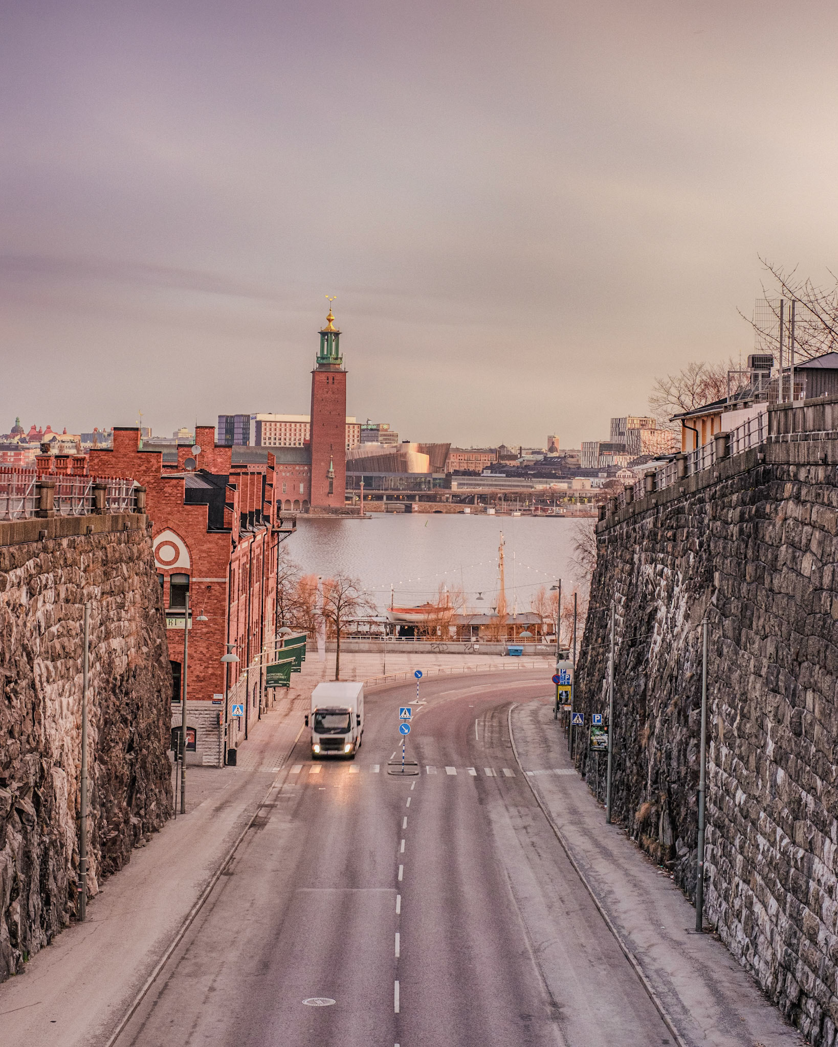 An empty dawn road winds between stone cliffs toward Riddarfjärden as Stockholm City Hall’s tower, topped by the golden Three Crowns, rises across the water.