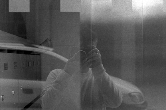 Autorretrato, fotografía analógica digitalizada, se ve a una fotografa sacando una foto a su propio reflejo en el cristal de un portal.