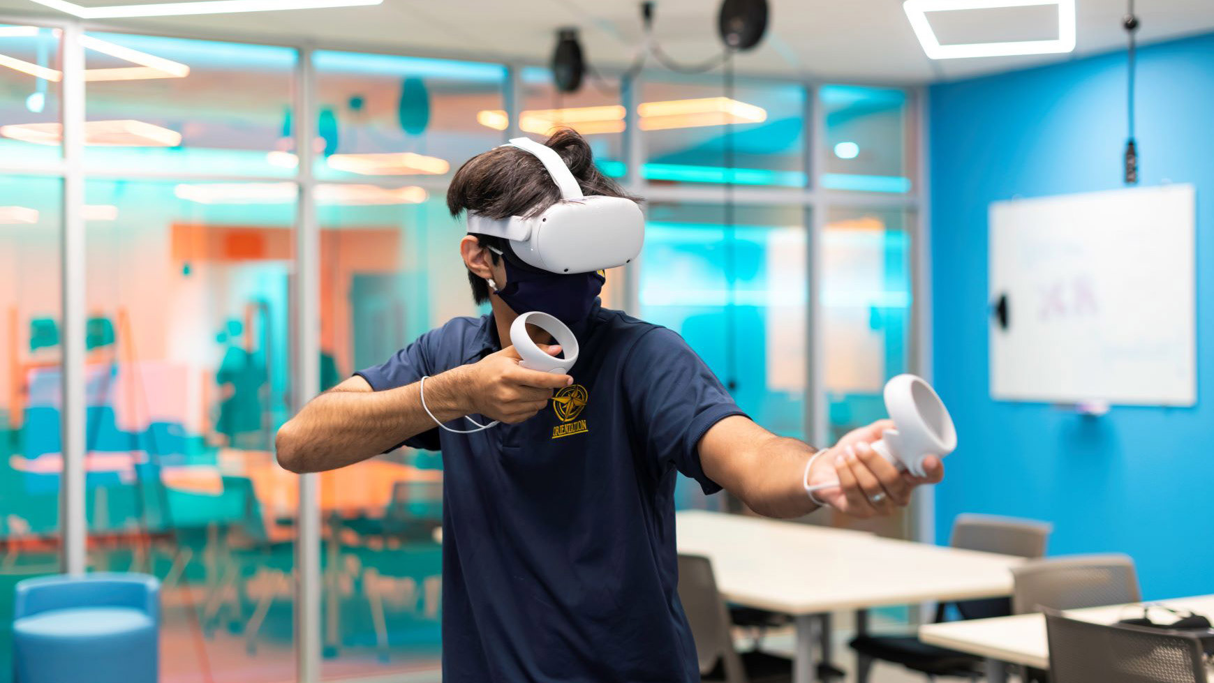 College boy with tan skin, brown hair, and navy t-shirt uses an oculus VR headset in front of Studio X's glass windows and blue walls.
