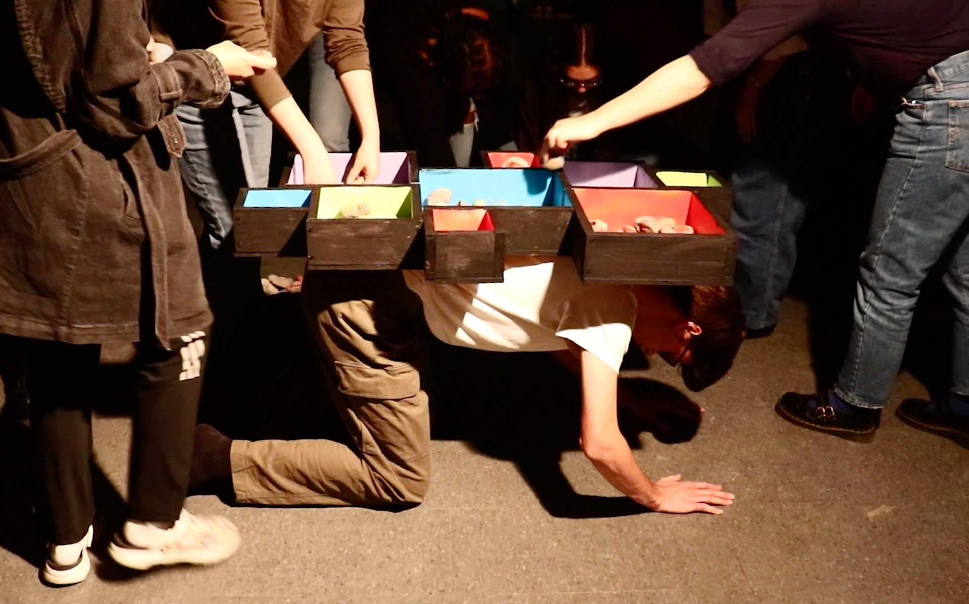 Performance video screenshot: the box structure is on my back, I'm in a table position on the floor, about 10 people surround me sorting rocks into the box.