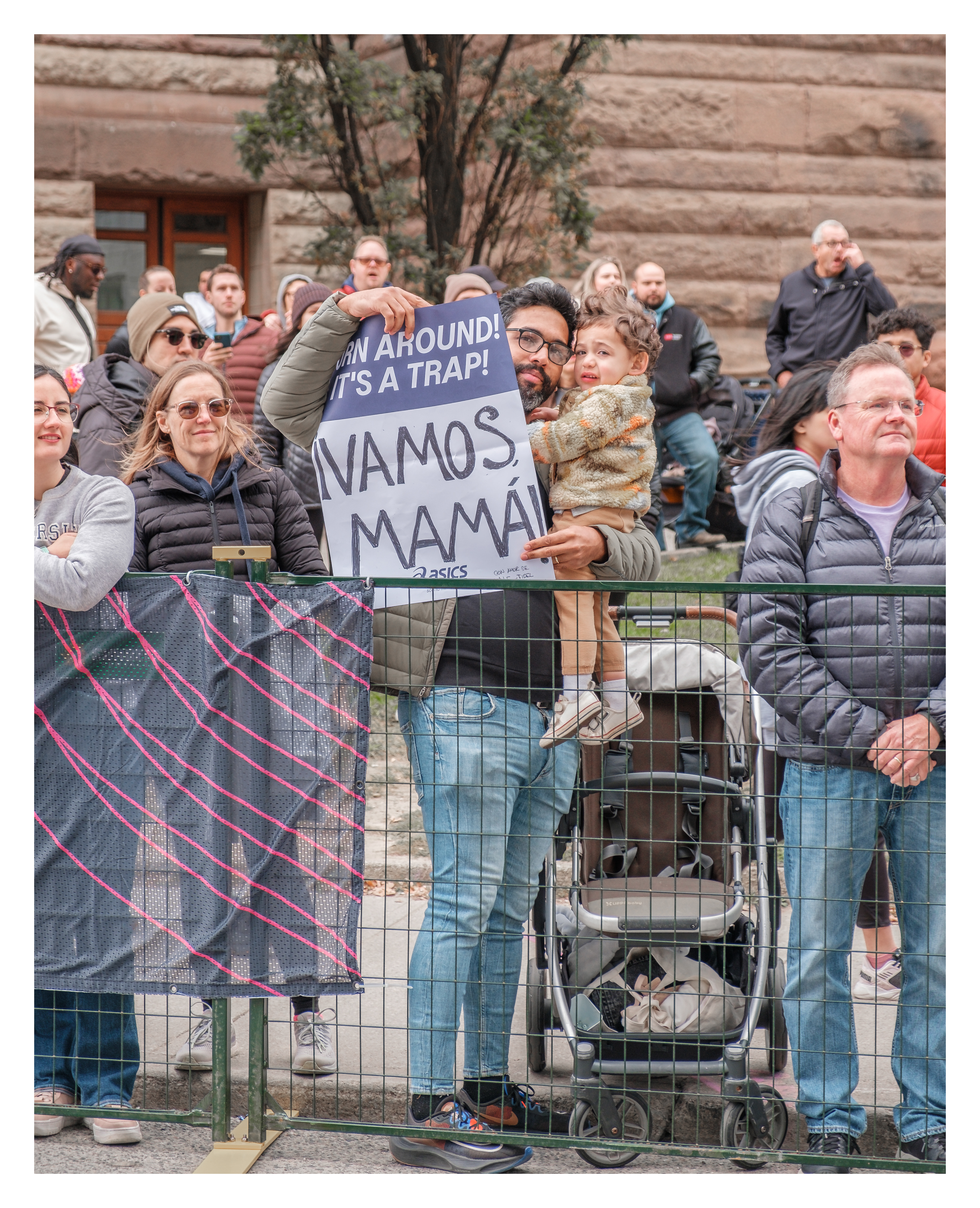 Man with child and crowd holding signs like “¡VAMOS MAMÁ!” during Toronto marathon, joyful multicultural support scene in urban athletic event.
