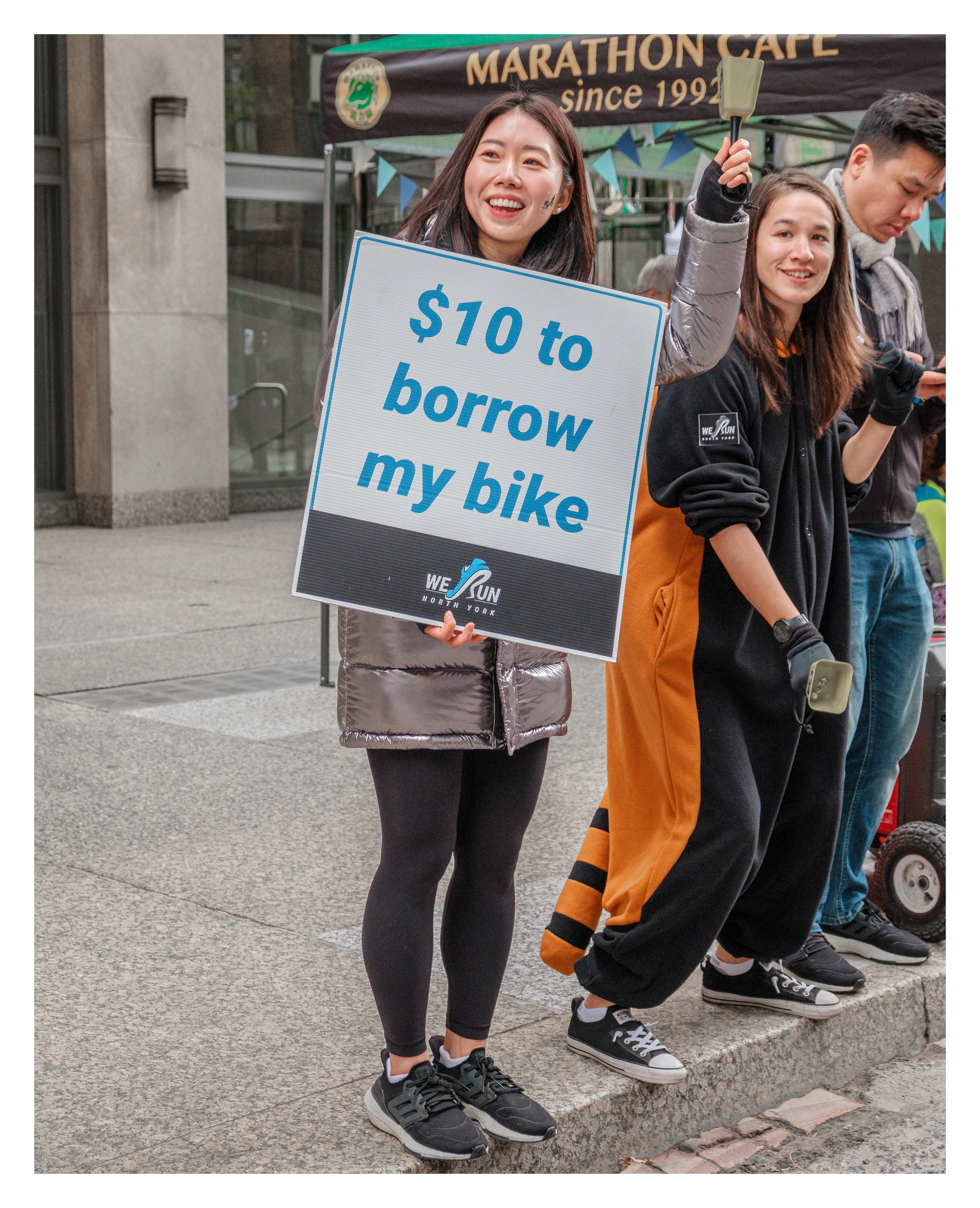 Group offering a humorous bike rental sign outside Marathon Café in Toronto, engaging passersby during a running event.