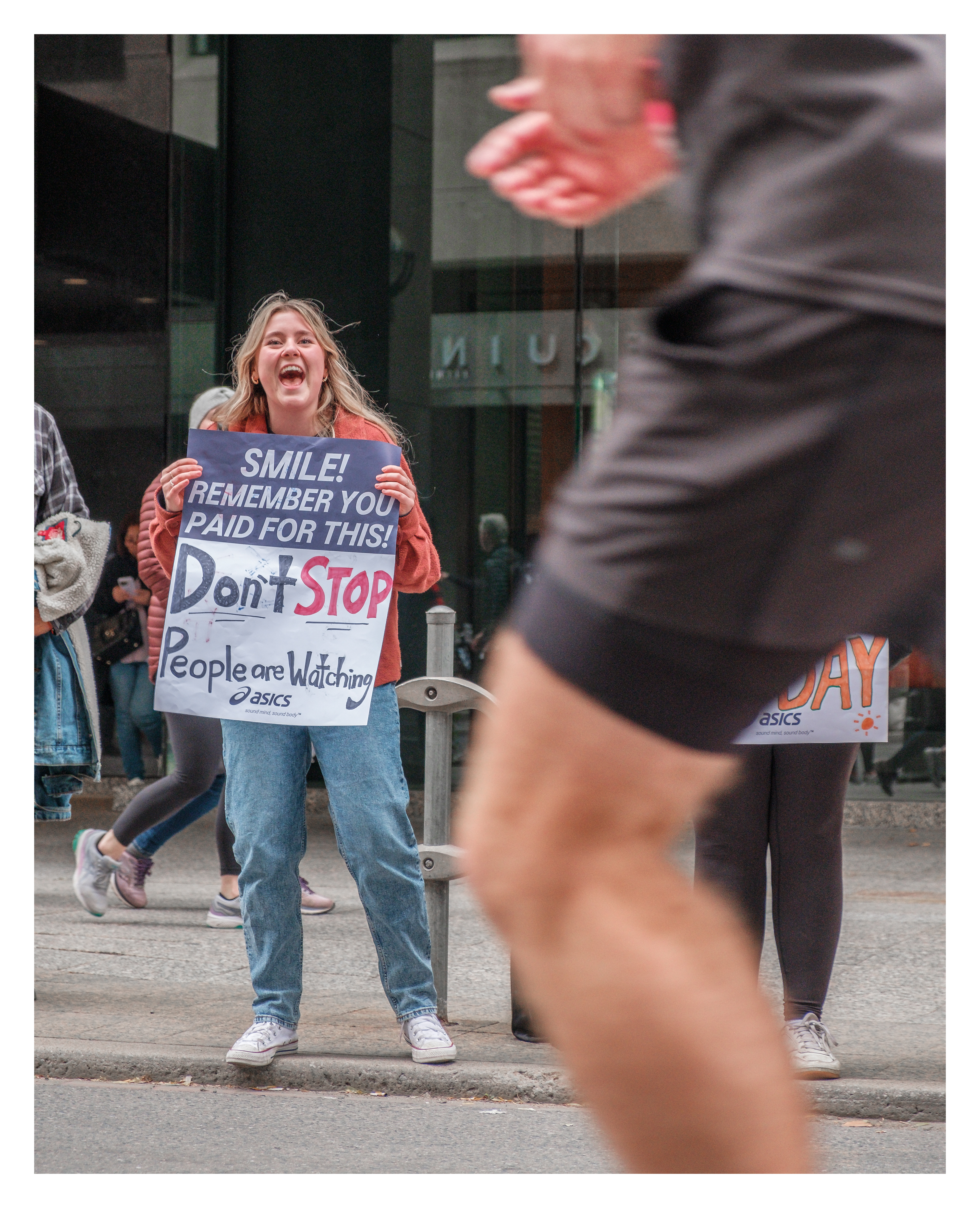 Marathon runner in motion passing woman with humorous sign: “Smile! Remember you paid for this”, street photography from the Toronto marathon finish area.