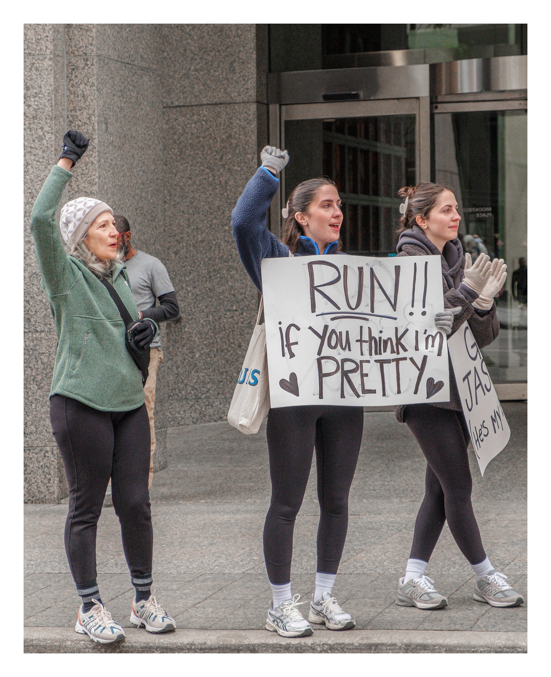 Three women holding a playful “Run if you think I’m pretty” sign in Toronto’s Financial District, expressing joy and encouragement during a city marathon.