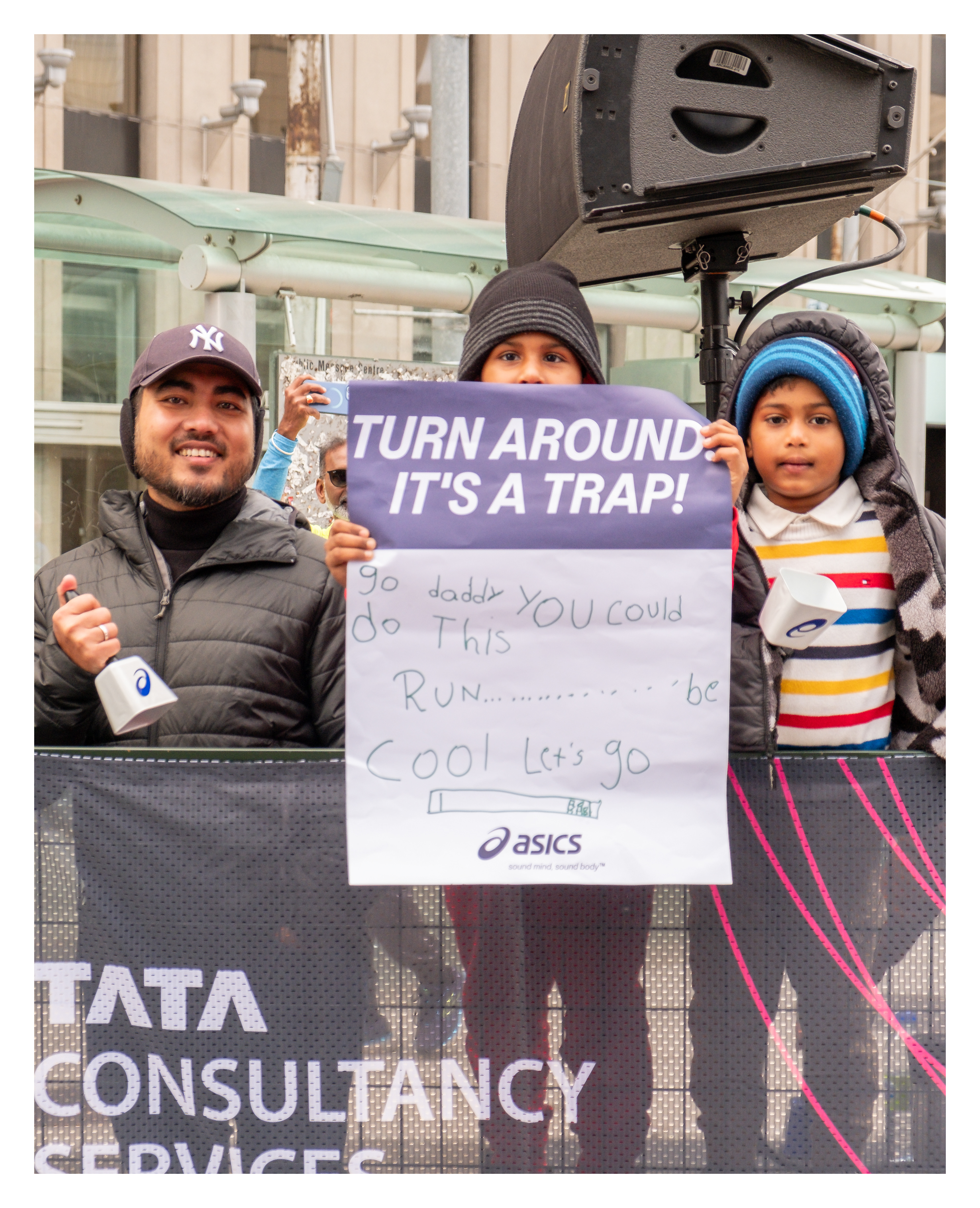 Toronto spectators holding humorous sign at marathon event near Queen Street, with children and adults cheering runners beside branded sponsor banners.