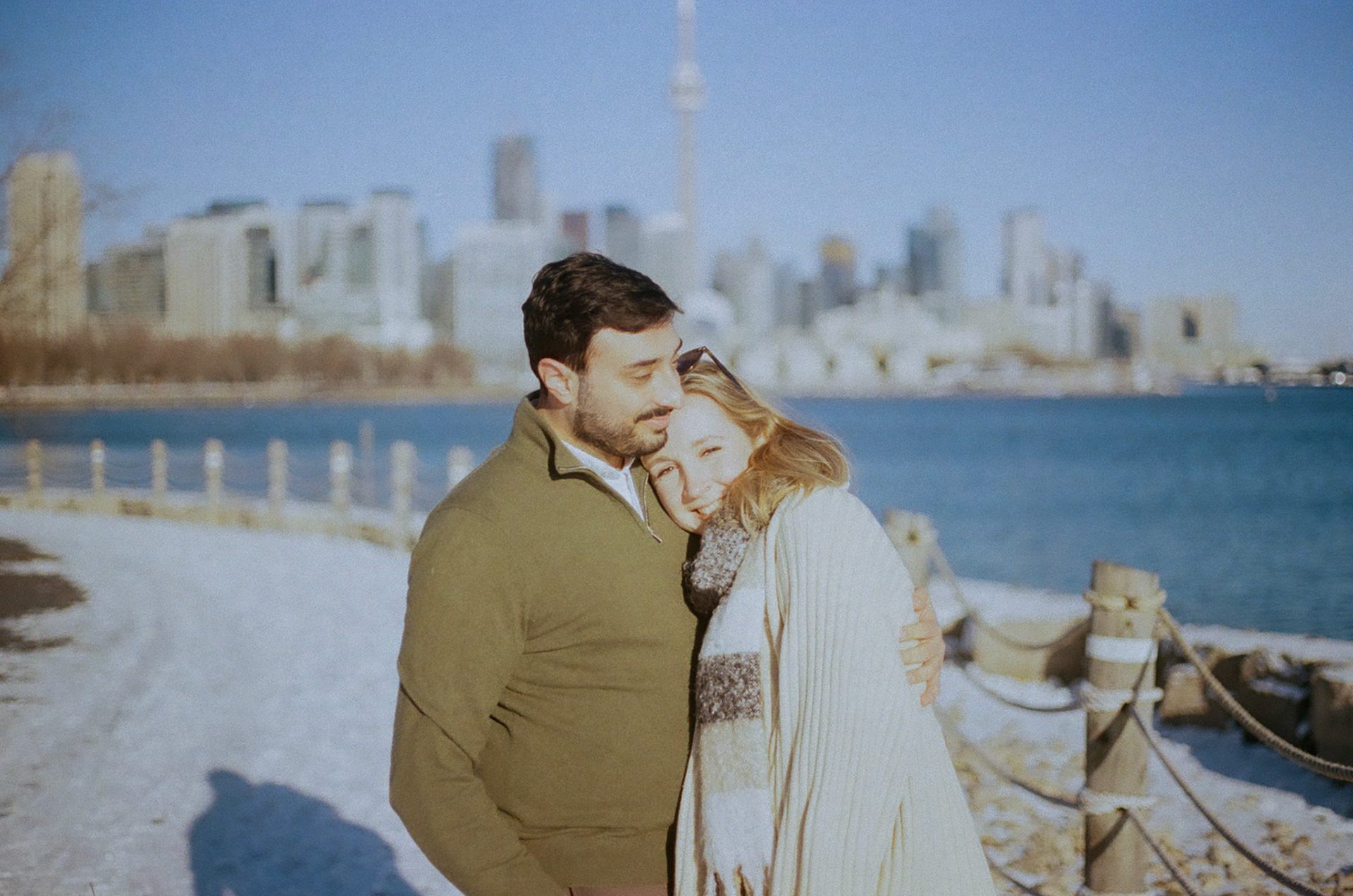 Man and woman walking side by side along Toronto waterfront, engaged and smiling under winter sun