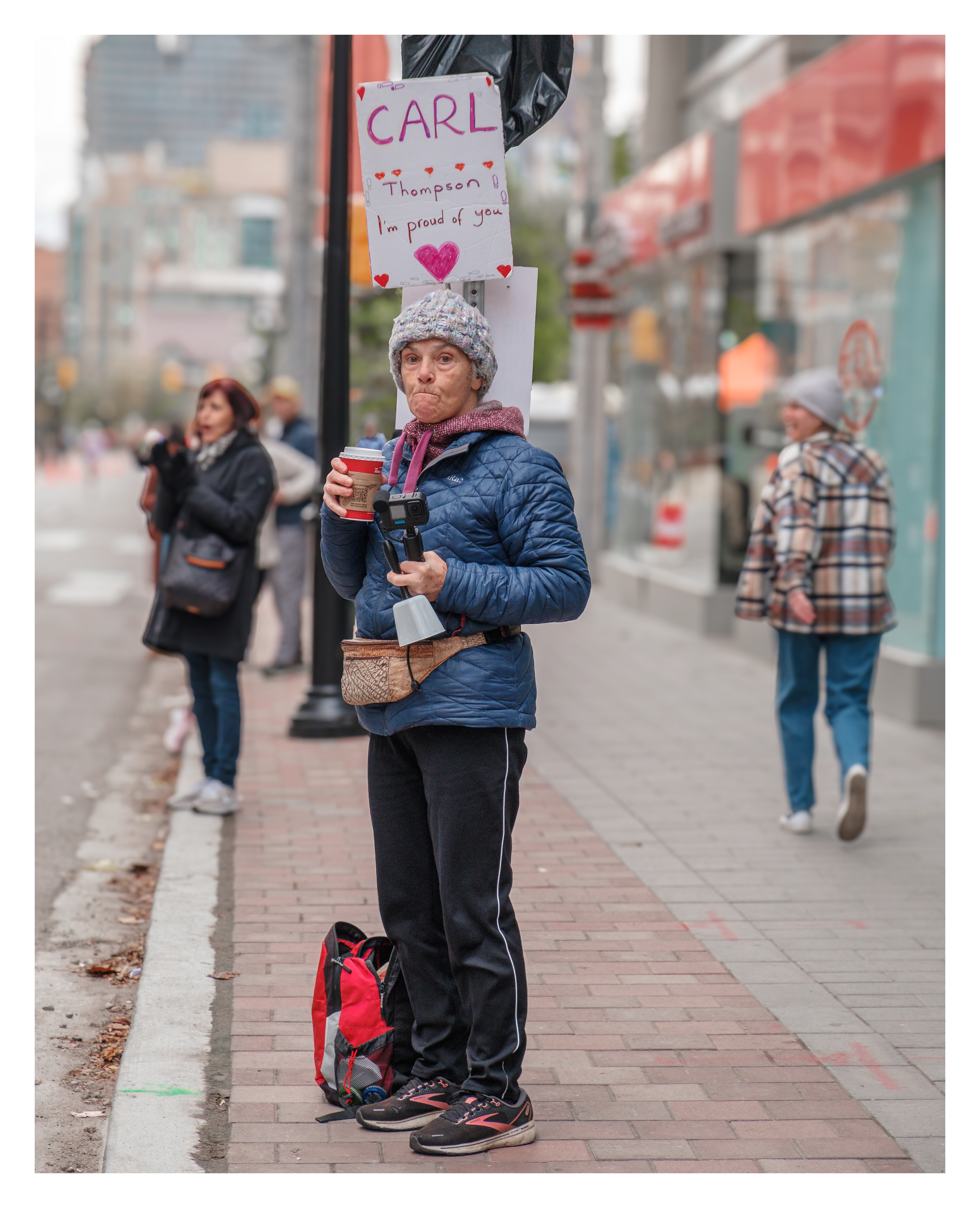 Woman with coffee cup cheering solo for runner named Carl in downtown Toronto, holding a personalized sign near commercial buildings.
