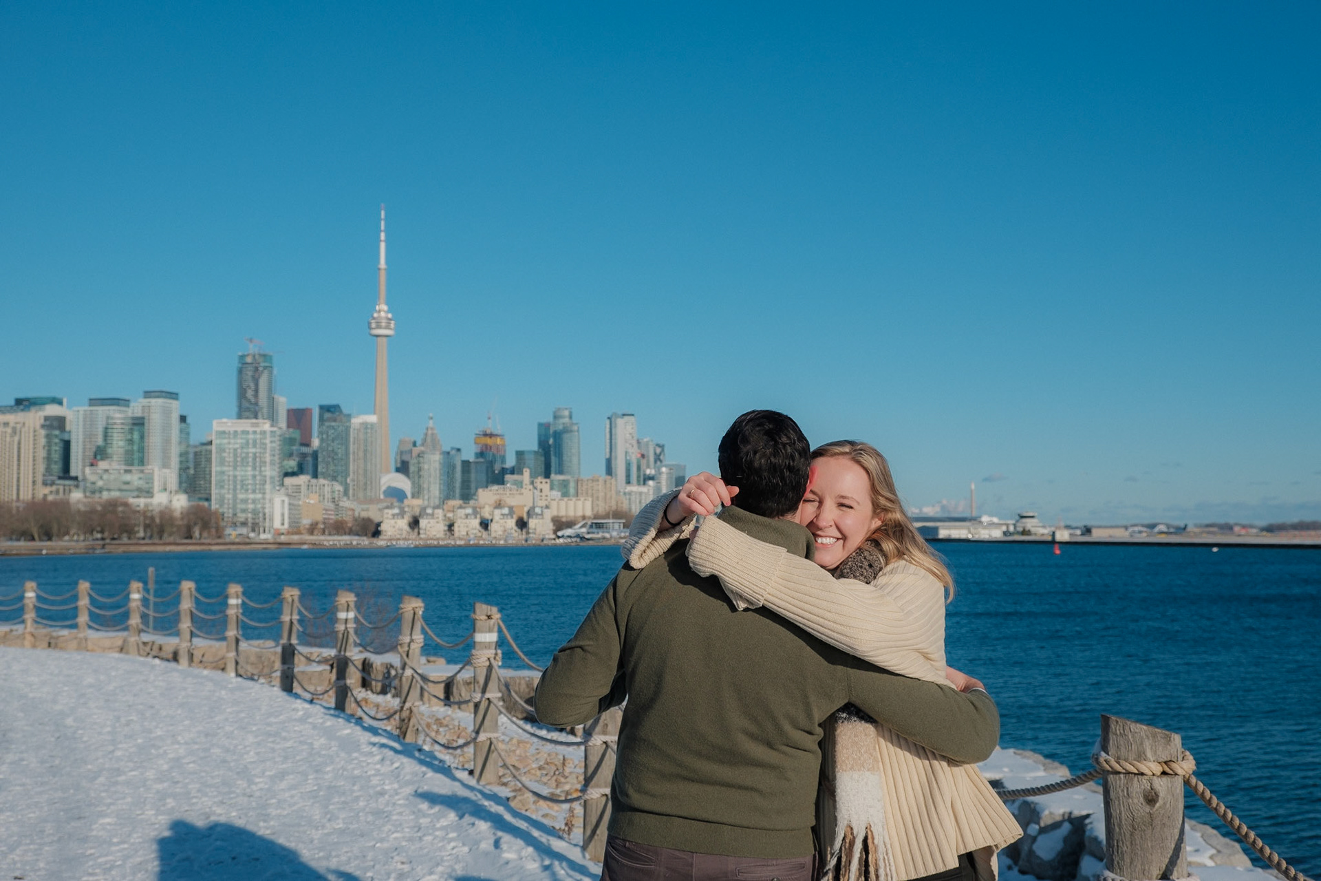 Engaged couple hugging with joyful expressions by the frozen lake, CN Tower in distant view