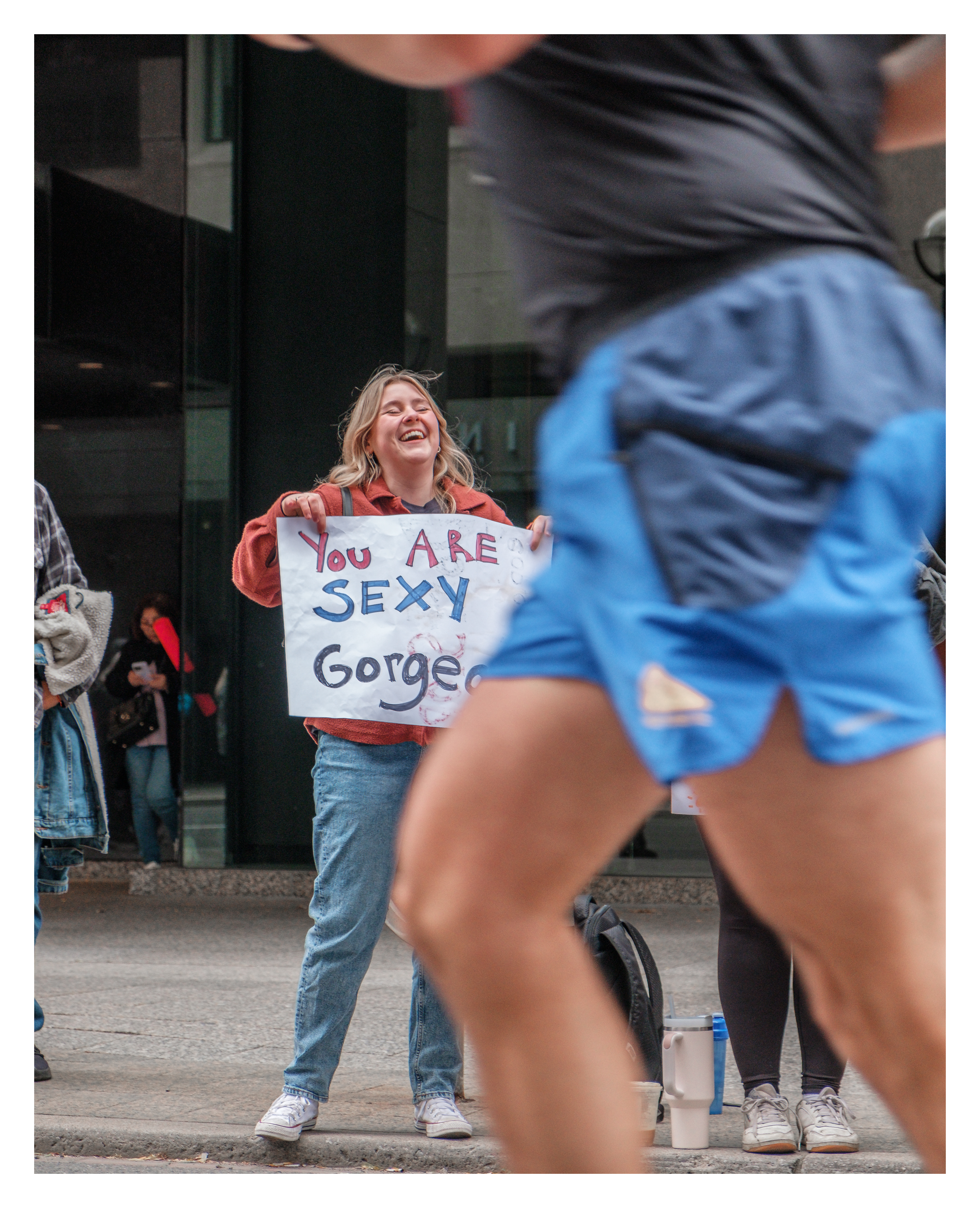 Candid moment of a runner passing a laughing woman holding sign reading “You are sexy, gorgeous”, capturing raw emotion in Toronto marathon street photography.