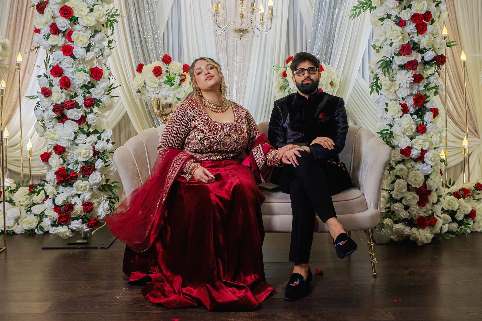 Couple seated on floral stage at South Asian engagement celebration in Toronto, with luxurious red and white floral backdrop