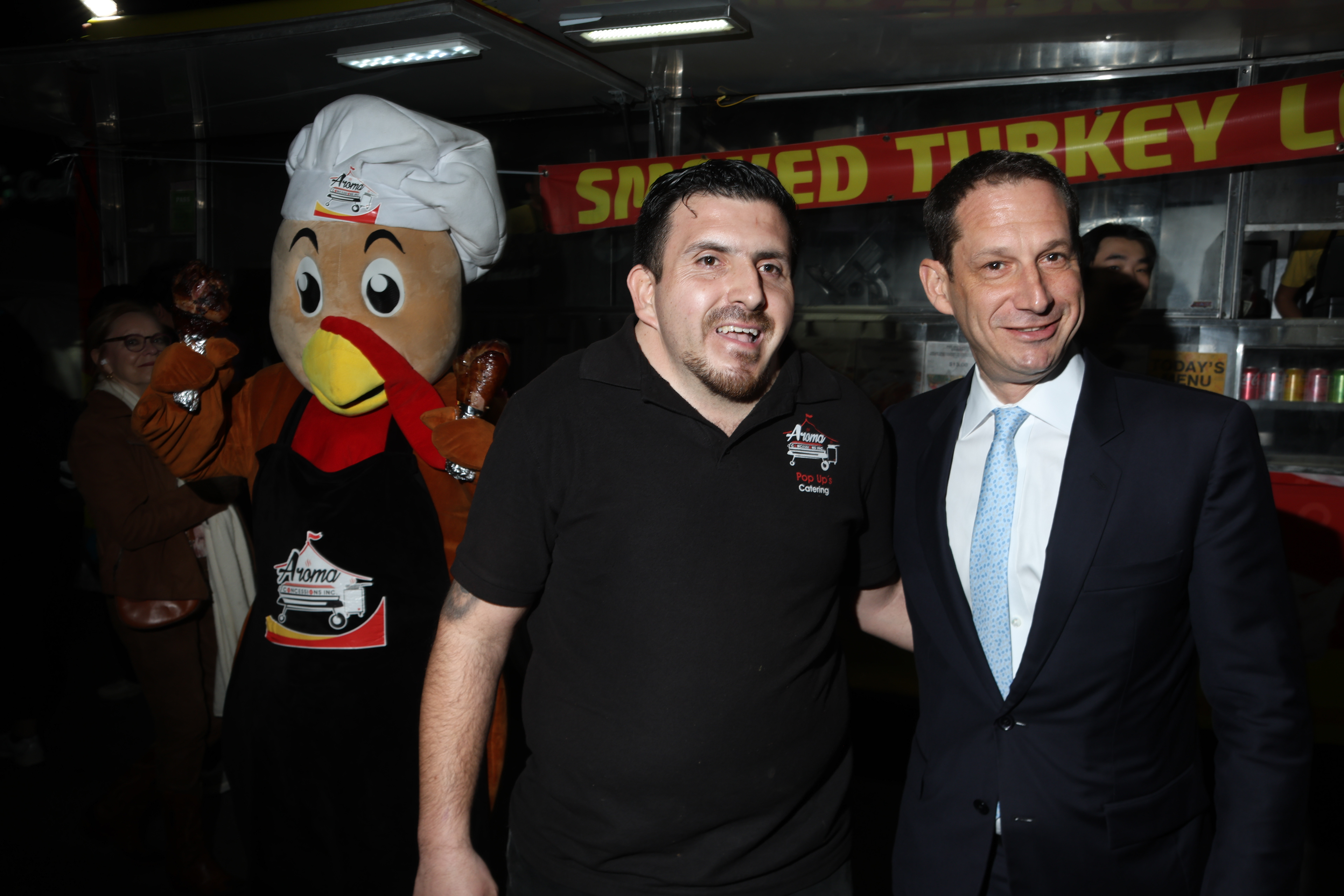 San Francisco Mayor Daniel Lurie smiles with one of the owners of Aroma Concessions, while mascot "Chef Turkey" lurks behind the pair during the Sunset Night Market on Februaru 27, 2026. Photo by Klyde Java.