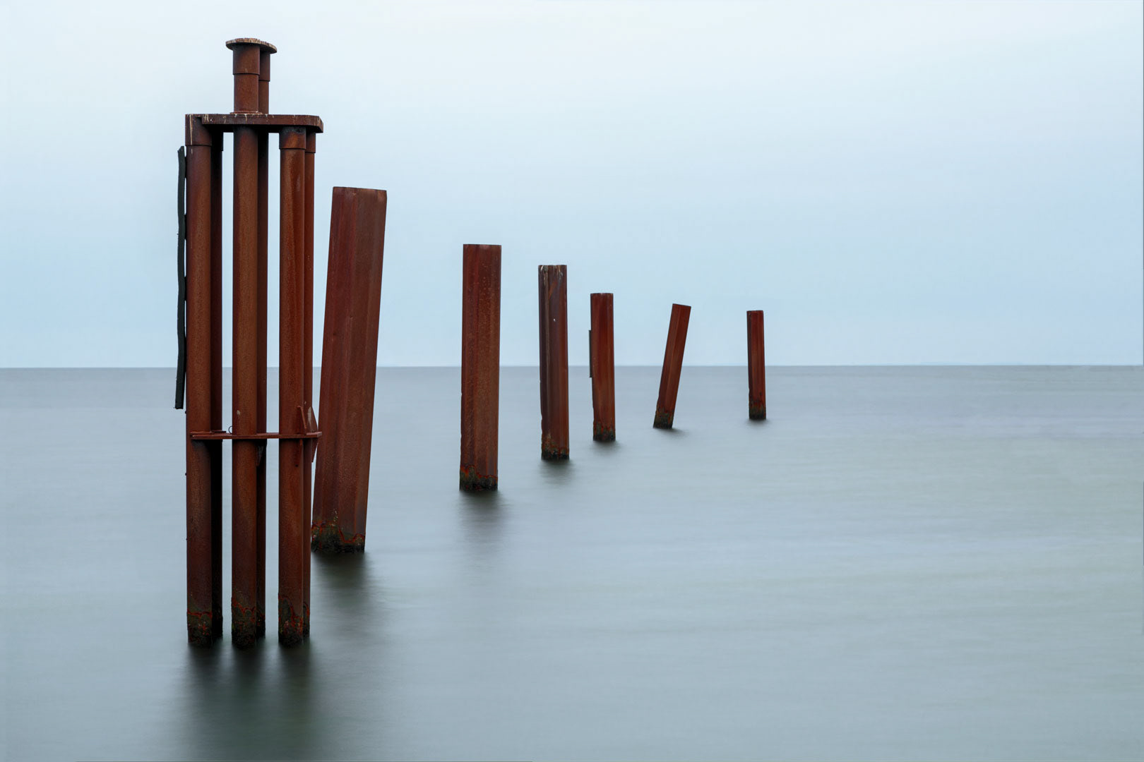 Minimal seascape photograph of rust-colored steel pillars emerging from calm water under a pale sky, expressing balance and stillness.