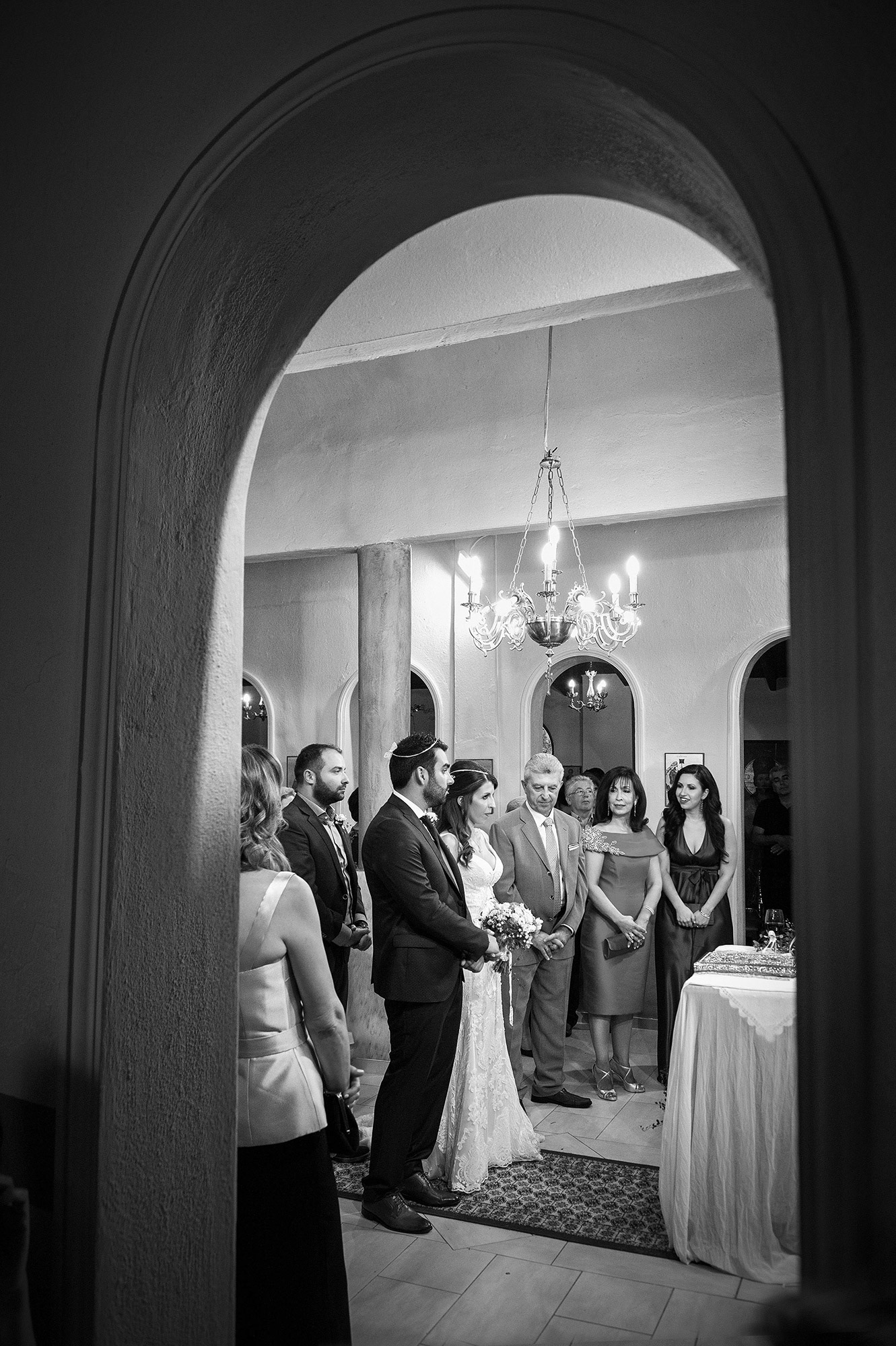 Το ζευγάρι κατά τη διάρκεια του μυστηρίου, φωτογραφημένο μέσα από την καμάρα της εκκλησίας σε ατμοσφαιρικό κάδρο.    The couple during the ceremony, captured through the church archway in a beautifully atmospheric frame.