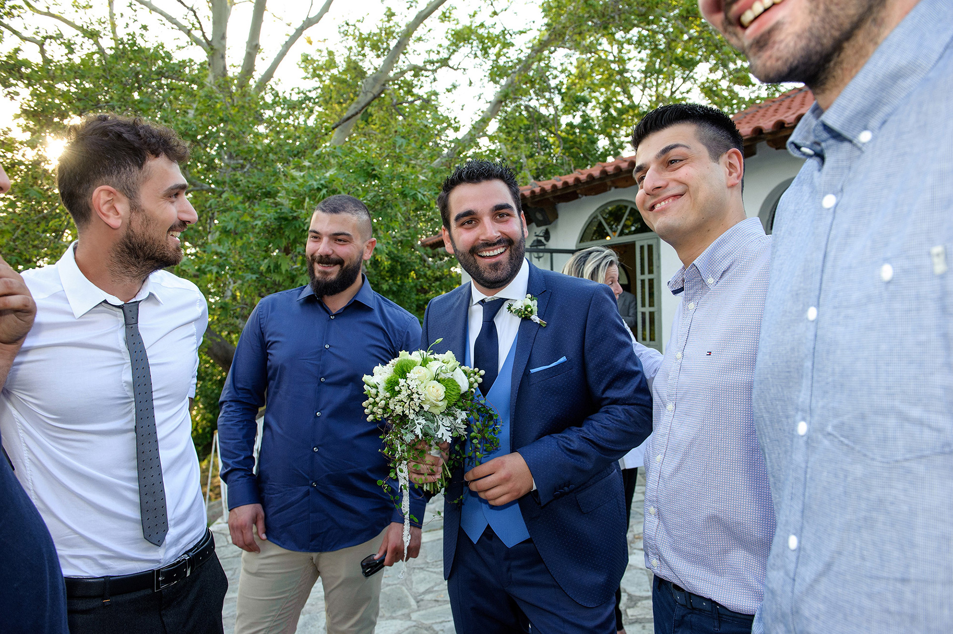 Ο γαμπρός με τους φίλους του έξω από την εκκλησία της Αγίας Τριάδας στη Θεσσαλονίκη, κρατώντας την ανθοδέσμη και χαμογελώντας.  The groom with his friends outside Agia Triada church in Thessaloniki, holding the bouquet and smiling.