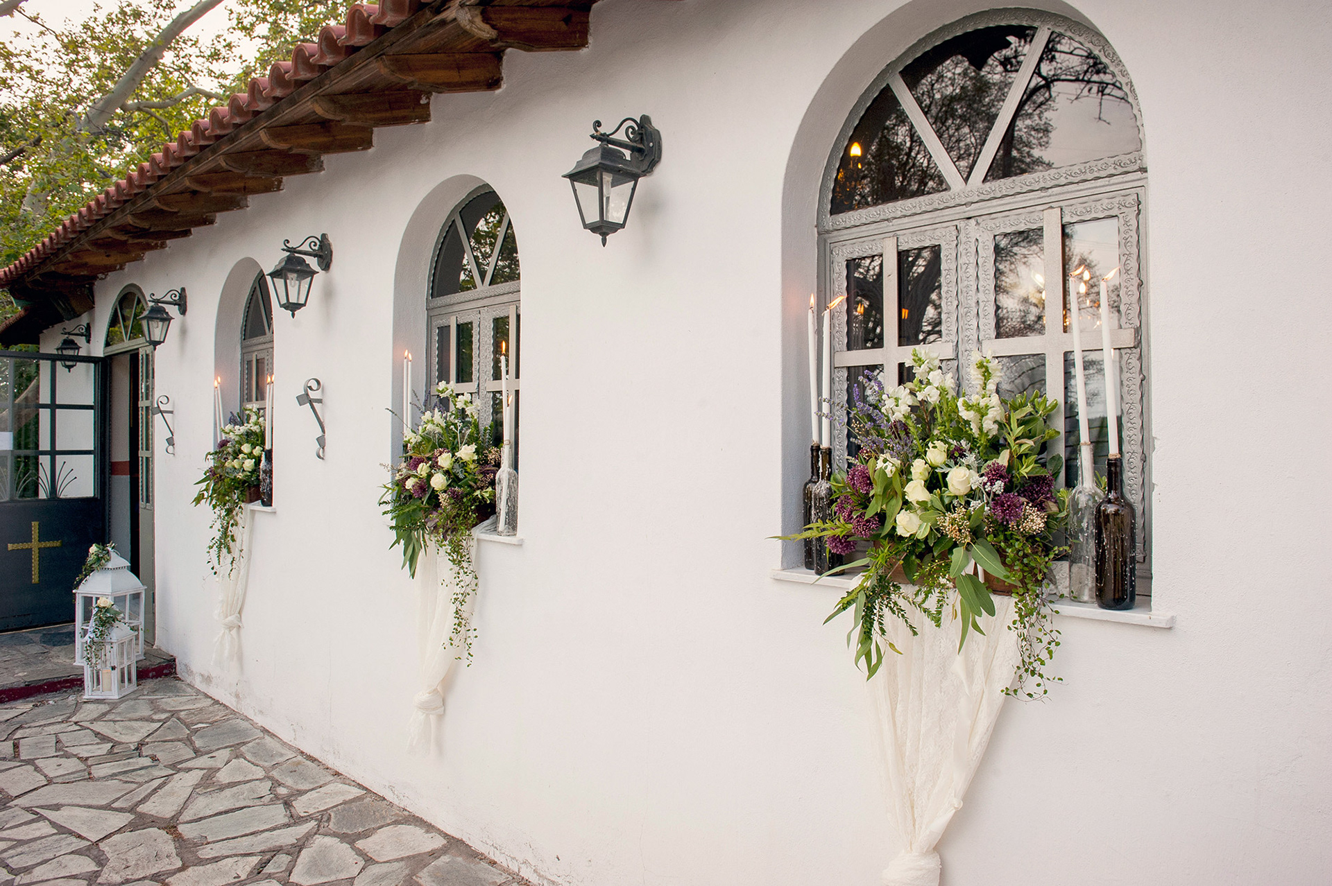 Εξωτερικός στολισμός εκκλησίας με λευκά και μοβ λουλούδια και κεριά στα παράθυρα.    Church exterior decorated with white and purple flowers and candles on the windows.