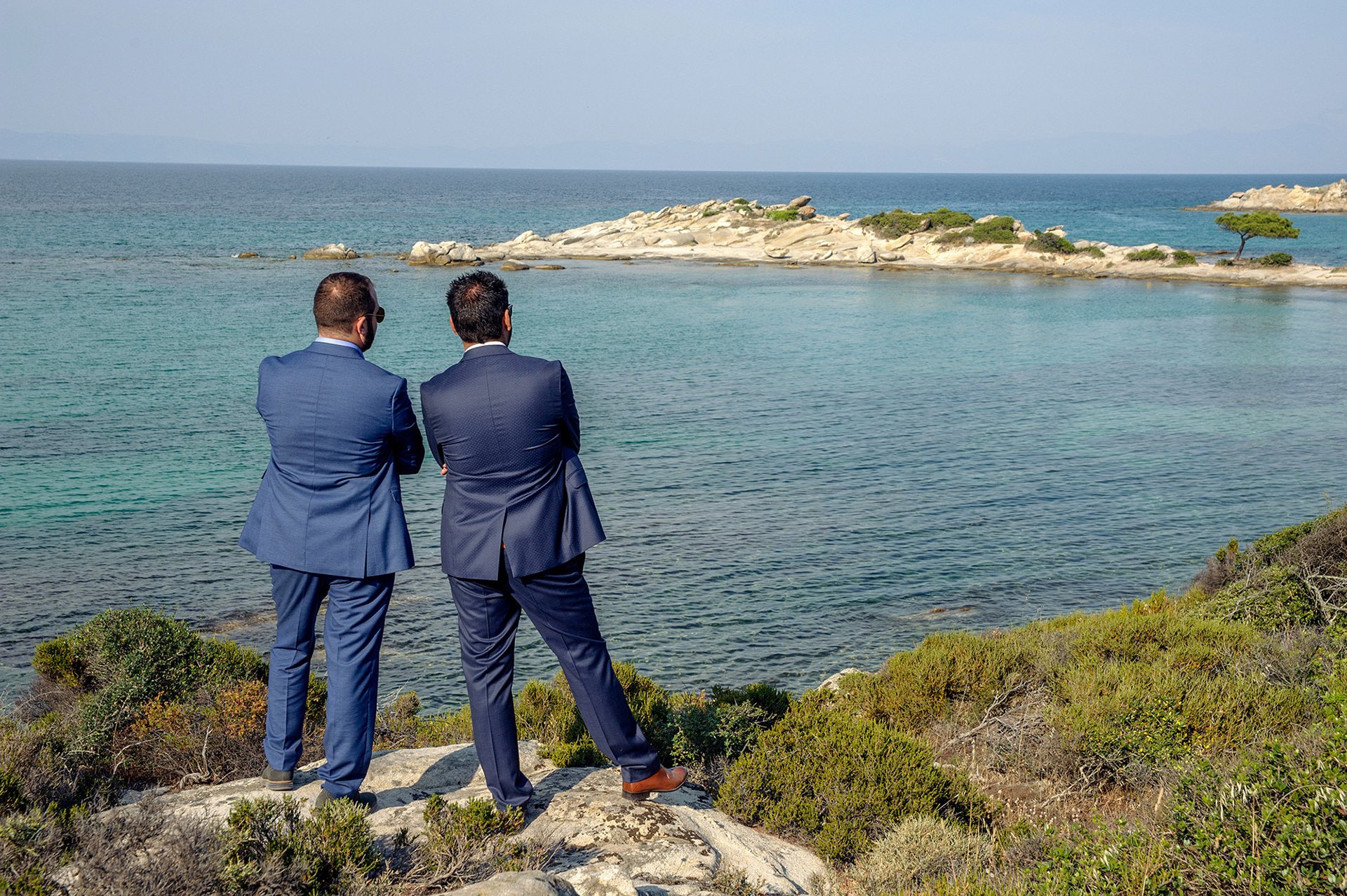 Ο γαμπρός με τον φίλο του στέκονται με πλάτη, κοιτάζοντας τη γαλάζια θάλασσα.    The groom and his friend stand side by side, looking at the blue sea.