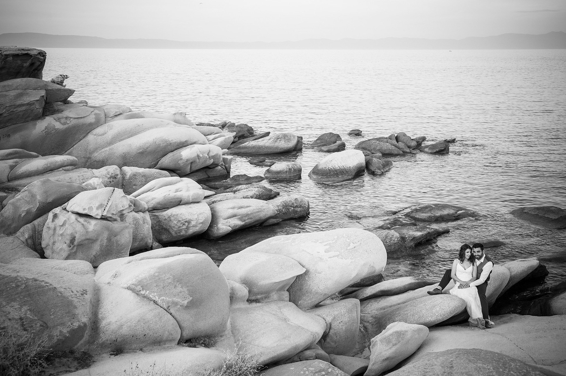 Το ζευγάρι κάθεται στα βράχια με φόντο τη θάλασσα της Χαλκιδικής, σε μαγευτική ασπρόμαυρη λήψη.    The couple sitting on the rocks with the Halkidiki sea behind them, stunning black and white wide shot.