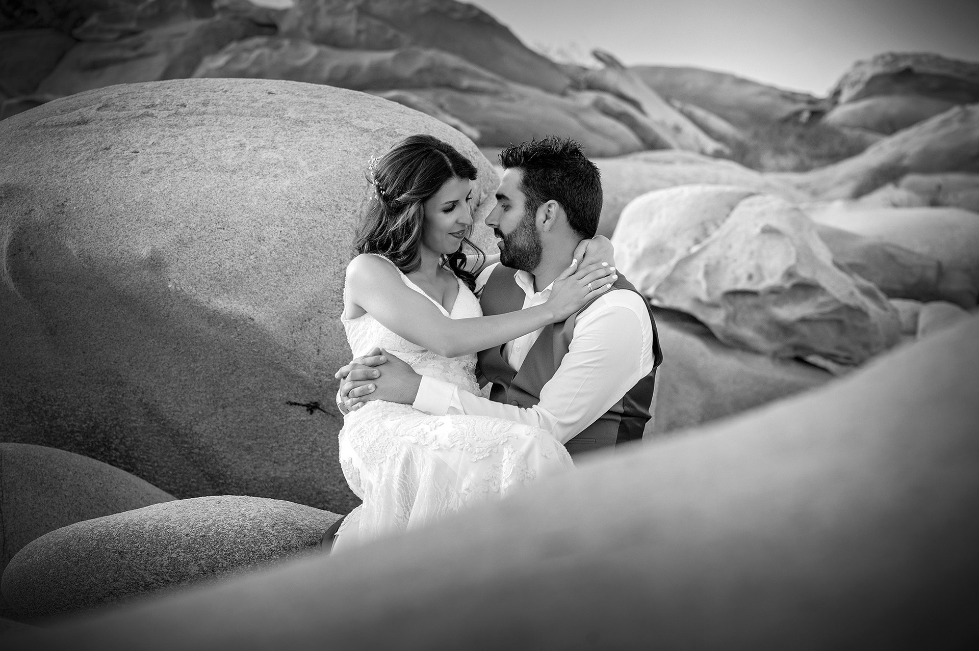 Ο γαμπρός και η νύφη αγκαλιάζονται τρυφερά ανάμεσα στα βράχια σε ρομαντική ασπρόμαυρη φωτογραφία.   The bride and groom embracing tenderly among the rocks in a romantic black and white shot.