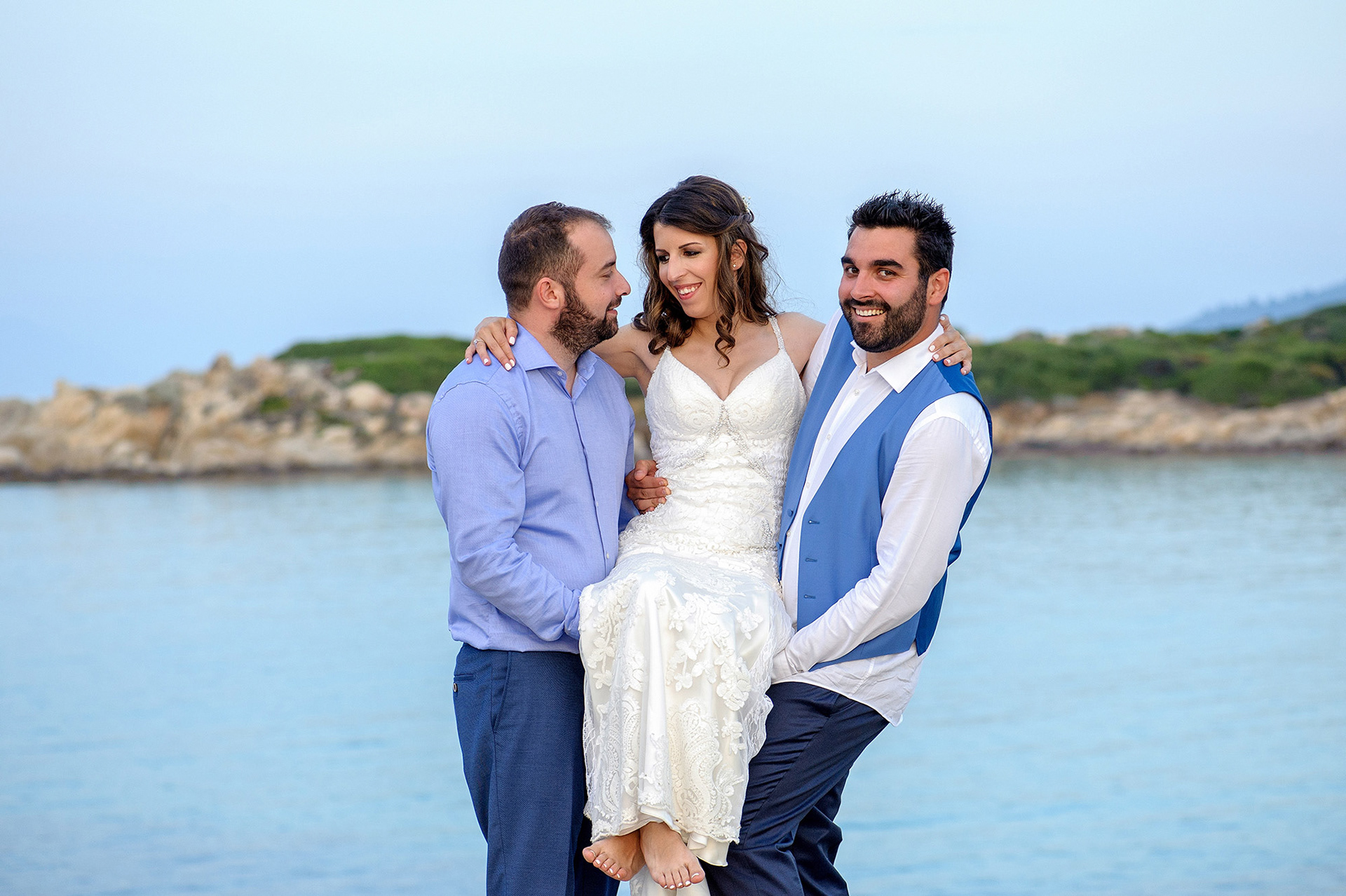 Η νύφη γελάει καθώς ο γαμπρός και ο φίλος της την κρατούν στα χέρια στην παραλία της Χαλκιδικής.    The bride laughing as the groom and a friend lift her in their arms on the beach in Halkidiki.