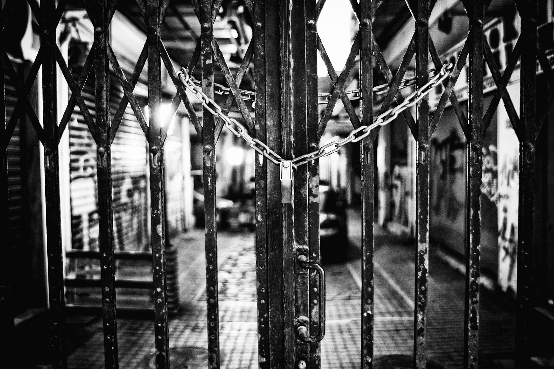Ασπρόμαυρη φωτογραφία αστικού τοπίου που εστιάζει σε μια κλειδωμένη μεταλλική πτυσσόμενη πόρτα με μια χοντρή αλυσίδα και λουκέτο, με φόντο μια σκοτεινή στοά της Θεσσαλονίκης, από τη φωτογράφο Βούλα Γκώτη.  Black and white urban photography focusing on a locked iron accordion gate with a heavy chain and padlock, framing a dark alleyway in Thessaloniki, shot by photographer Voula Gkoti.