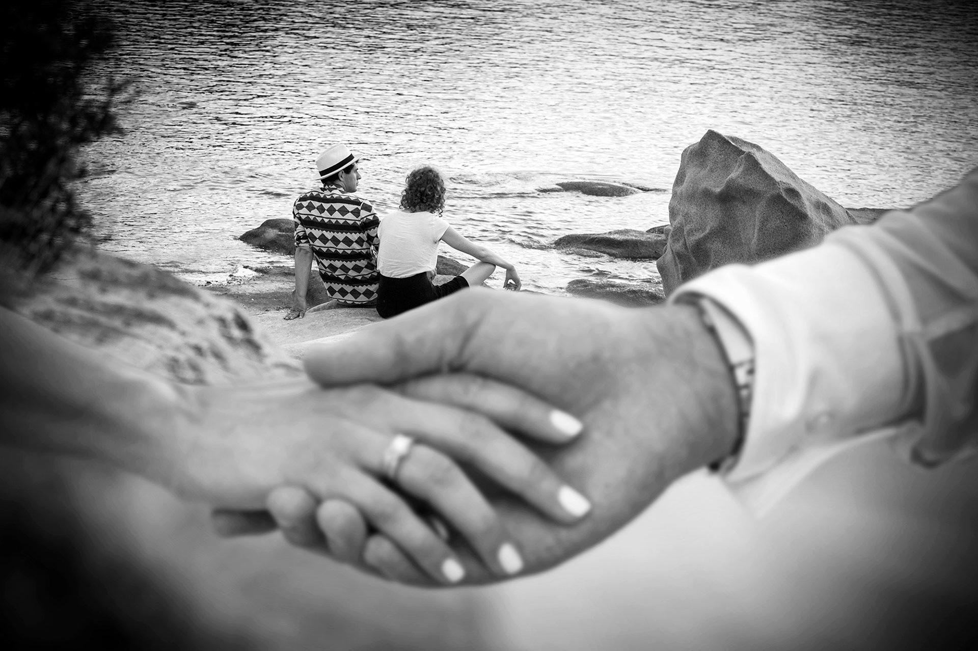Εστίαση στα ενωμένα χέρια του ζευγαριού, με φόντο τη θάλασσα και ένα ζευγάρι να κάθεται στα βράχια.    Focus on the couple’s joined hands, with another pair sitting by the sea in the background.