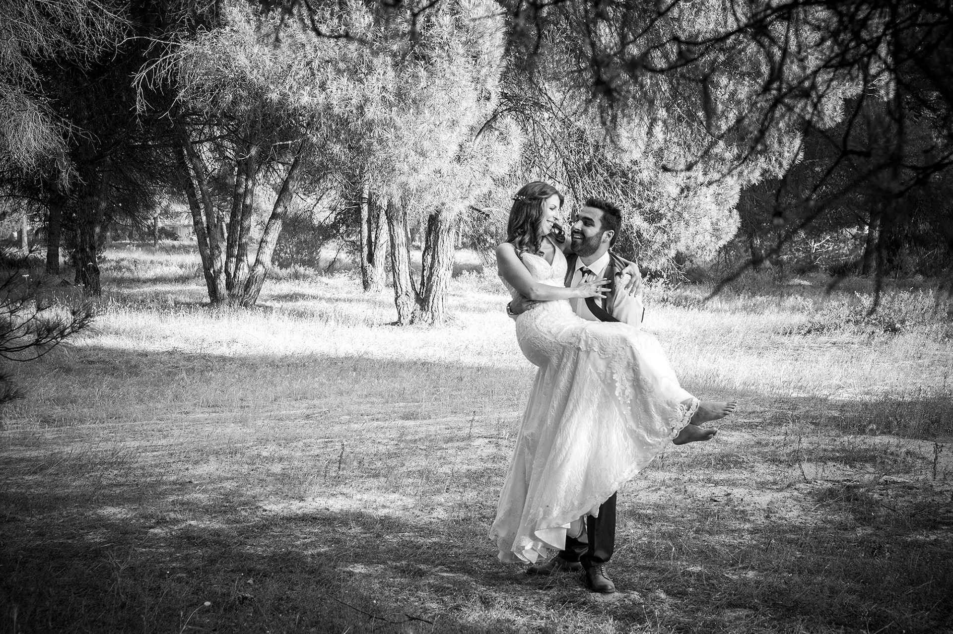 Μαυρόασπρη φωτογραφία όπου ο γαμπρός κρατάει στα χέρια του τη νύφη ανάμεσα στα δέντρα.    Black and white photo of the groom lifting the bride in his arms among the trees.