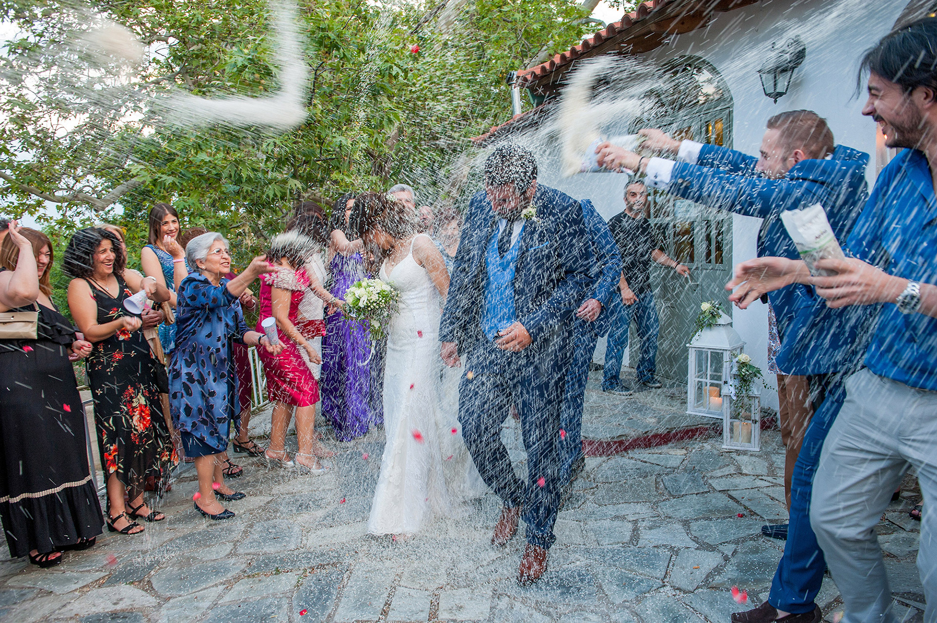 Το ζευγάρι γελάει και χαίρεται καθώς το ρύζι πετάγεται παντού, σε μια χαρούμενη στιγμή μετά το μυστήριο.   The couple laughs with joy as rice fills the air in a cheerful moment right after the ceremony.