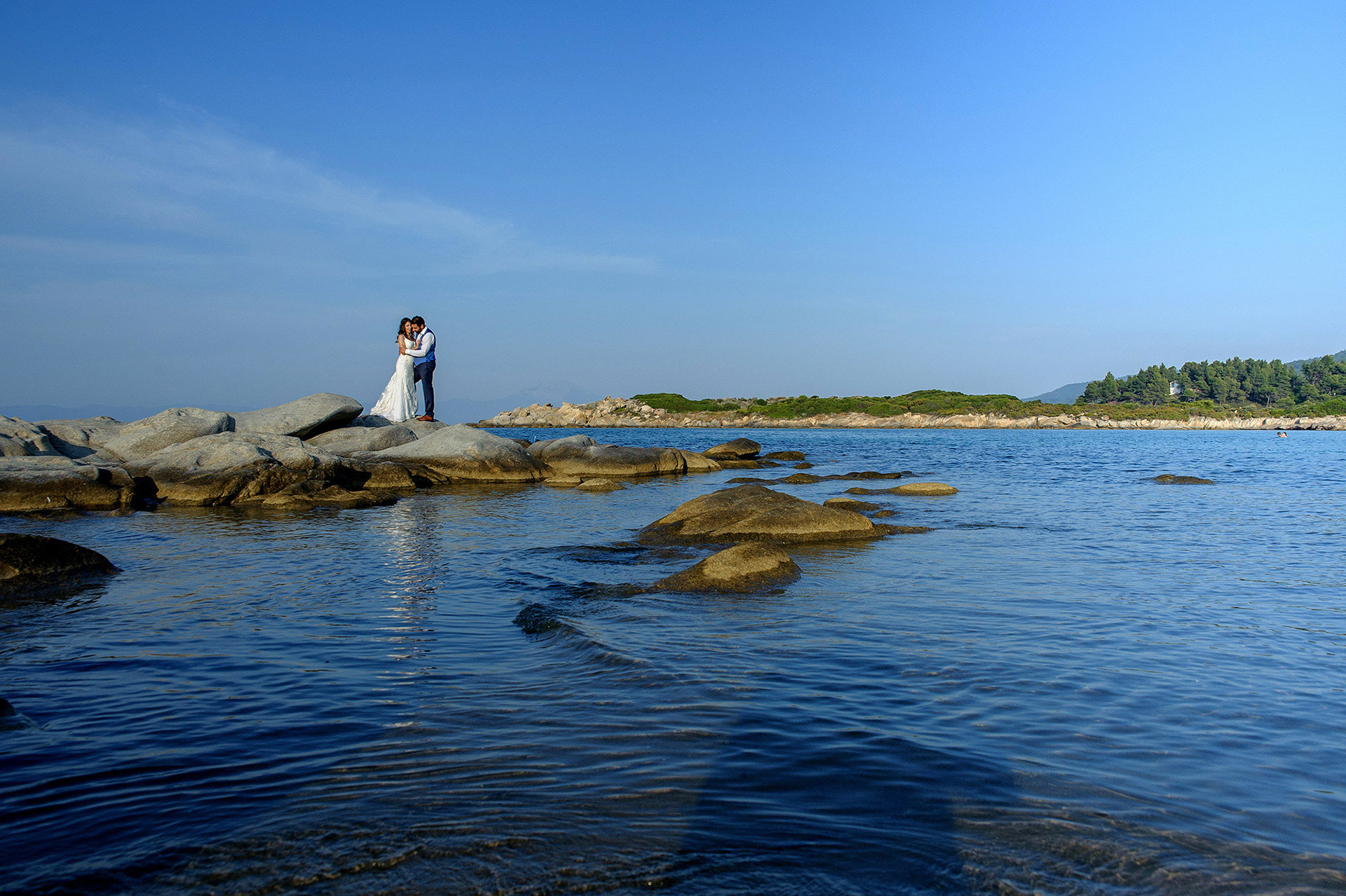 Το ζευγάρι αγκαλιάζεται πάνω στα βράχια με φόντο τη θάλασσα και το ηλιοβασίλεμα.   The couple embraces on the rocks by the sea with the sunset in the background.